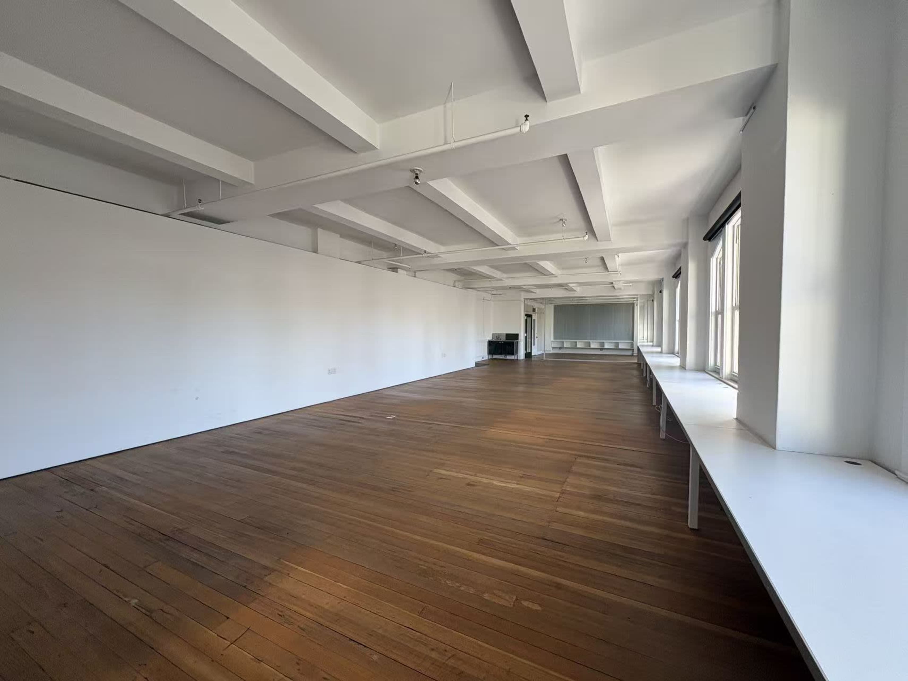 The image shows an empty room with wooden floors, white walls, and large windows along one side.