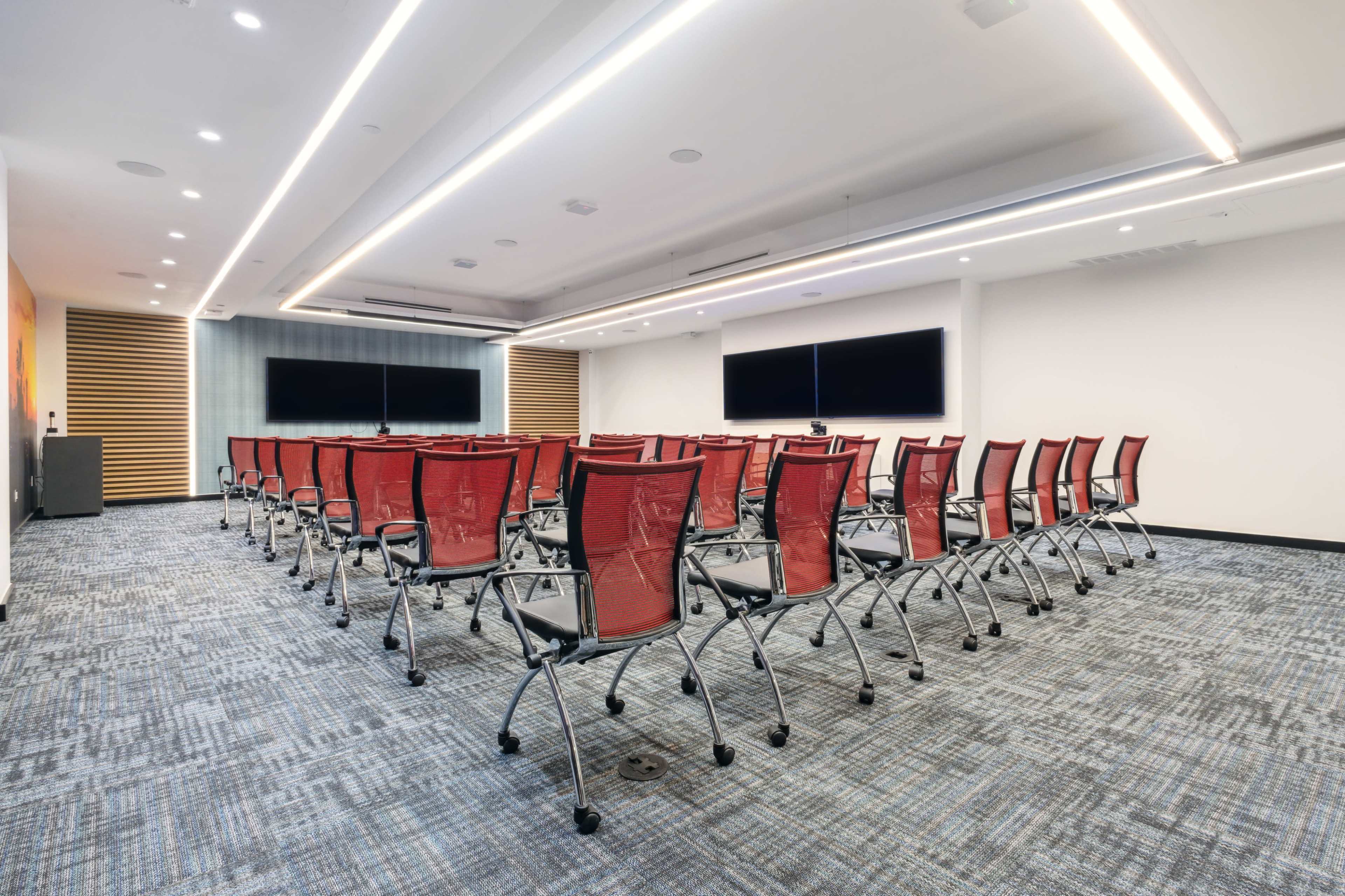 A modern conference room arranged with rows of red mesh chairs facing two large screens on the front wall.