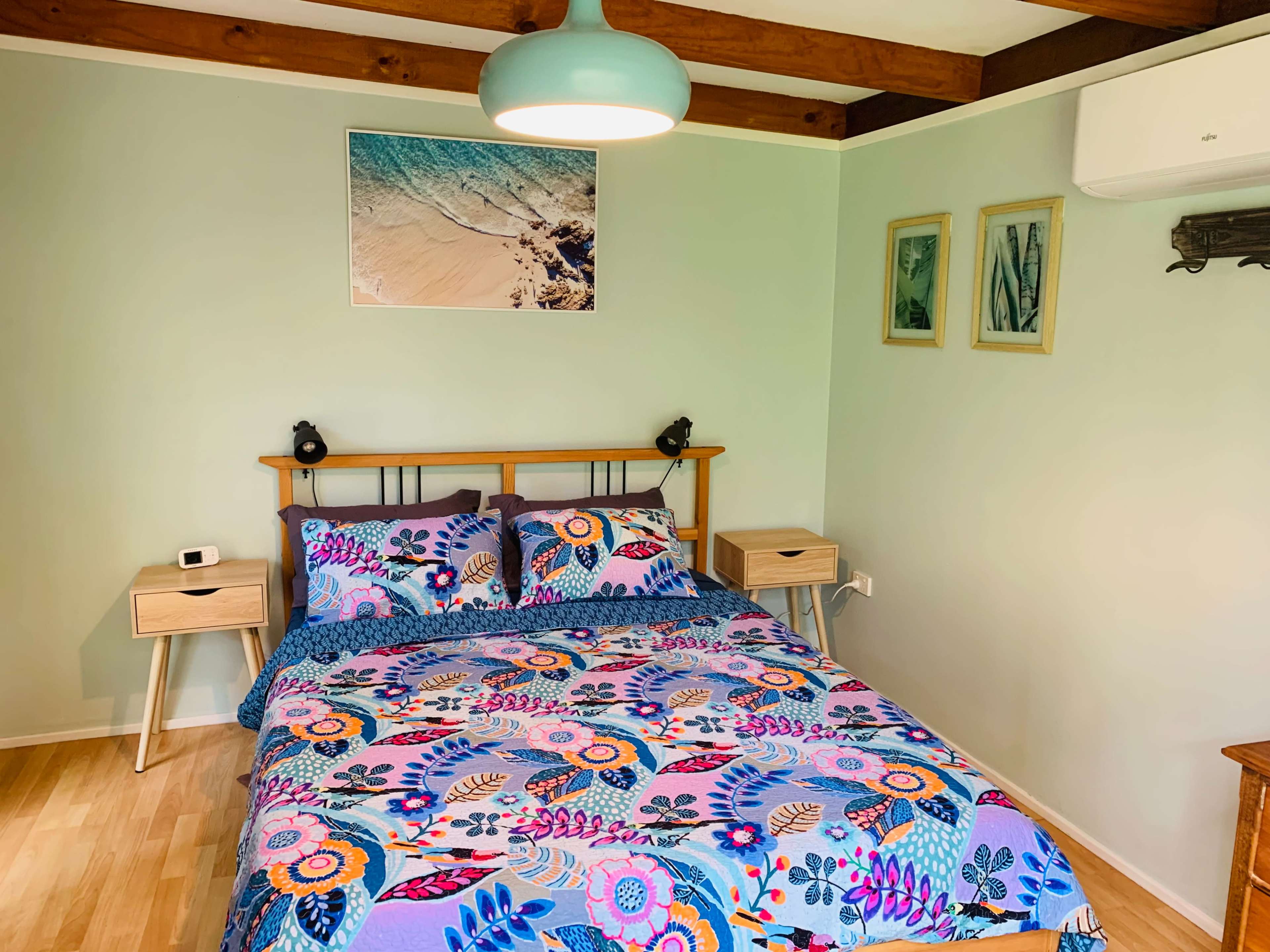 The image shows a brightly colored bedroom featuring a floral-patterned bedspread, wooden furniture with bedside tables, and a beach-themed artwork on the wall.