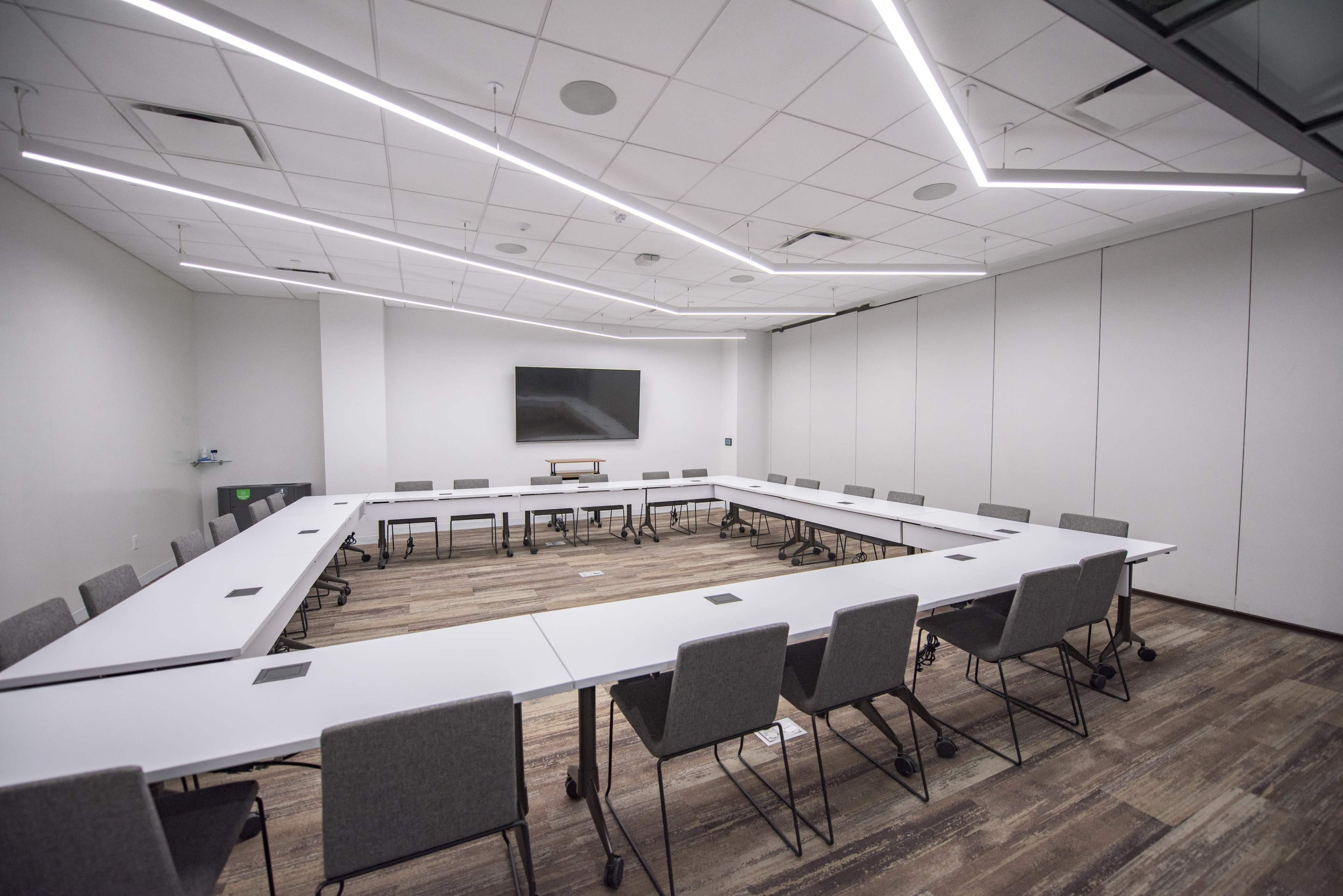 A modern conference room features a large rectangular table surrounded by gray chairs, with a wall-mounted screen and recessed lighting.