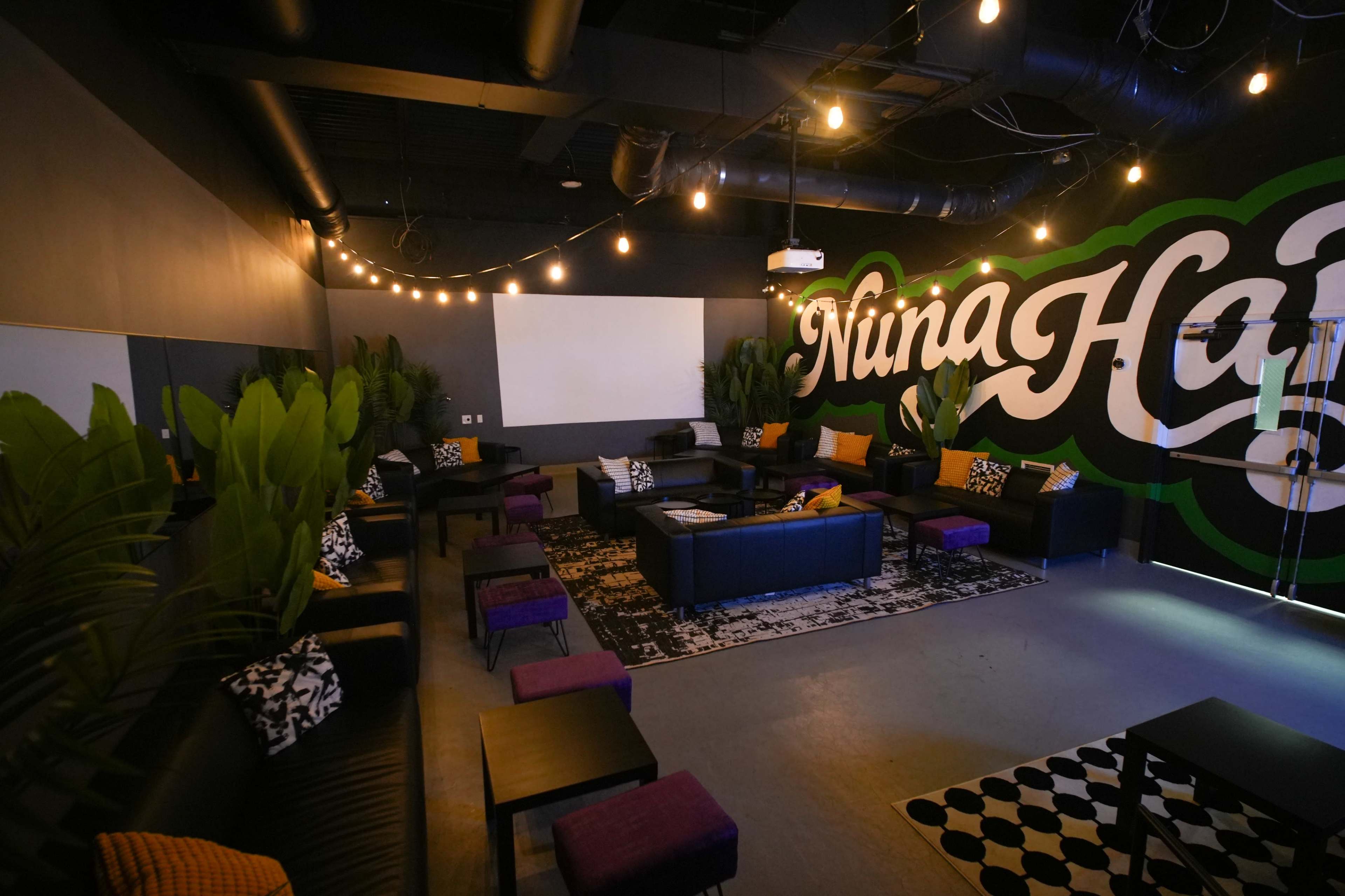 The image shows a modern lounge area with black leather sofas, decorative pillows, a patterned rug, and a large mural reading "Nina Ha." Soft lighting from string lights illuminates the room.