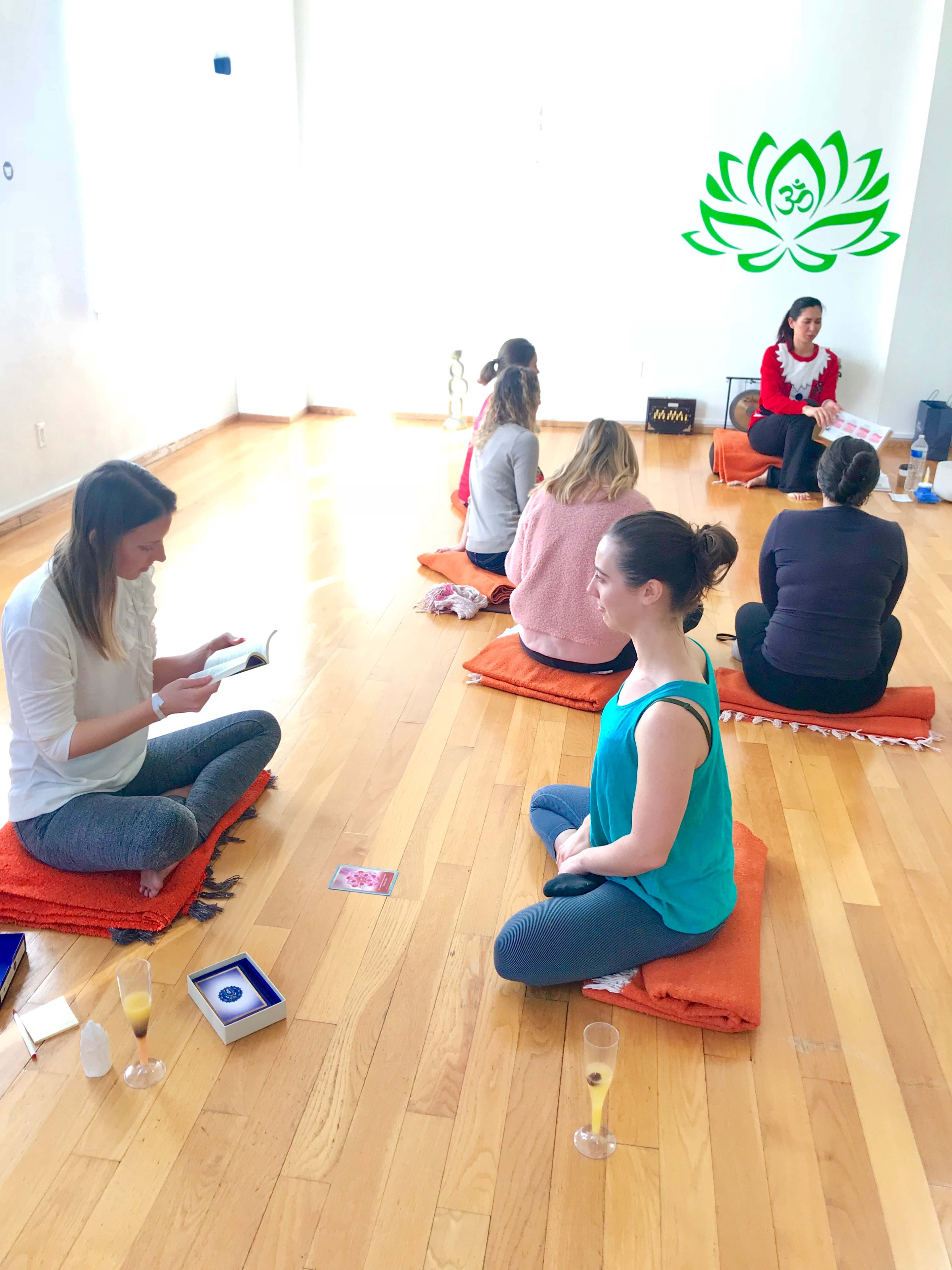 A group of people sits on orange mats in a bright room, engaged in a meditation session led by a woman at the front.