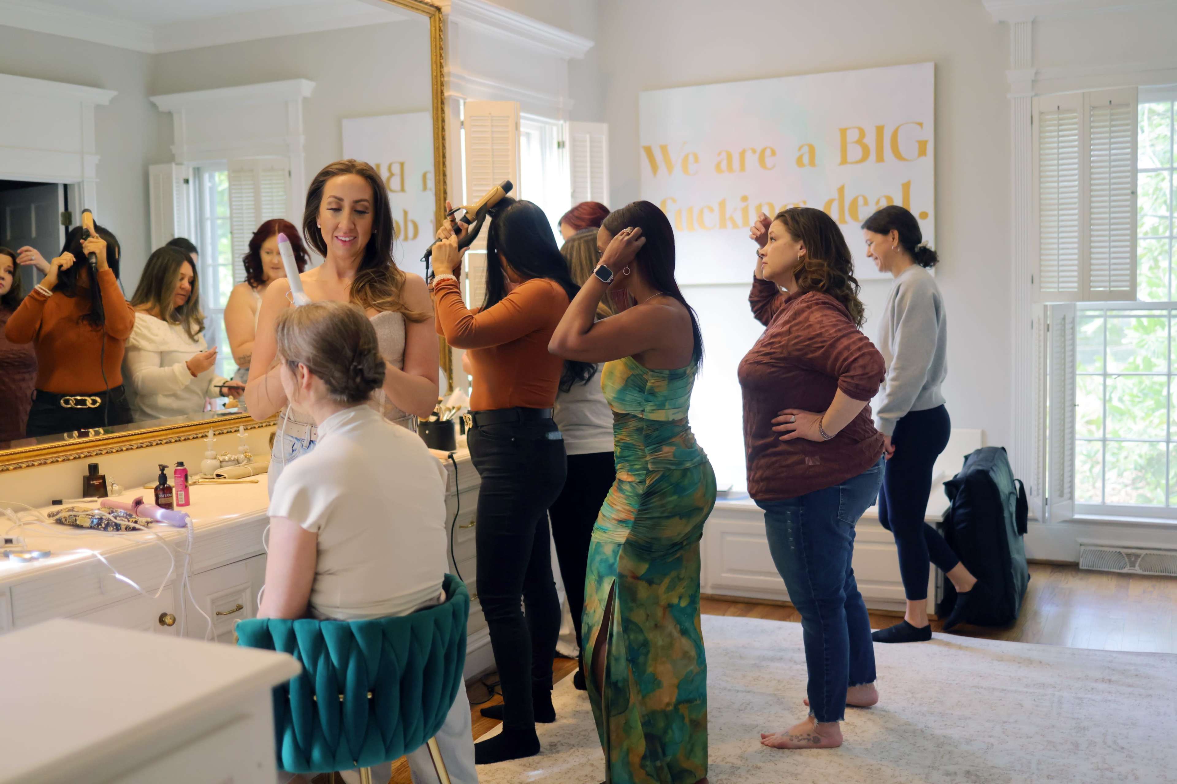 A group of women are preparing for an event in a well-lit room with a large mirror, where some are styling their hair and others are engaged in conversation.
