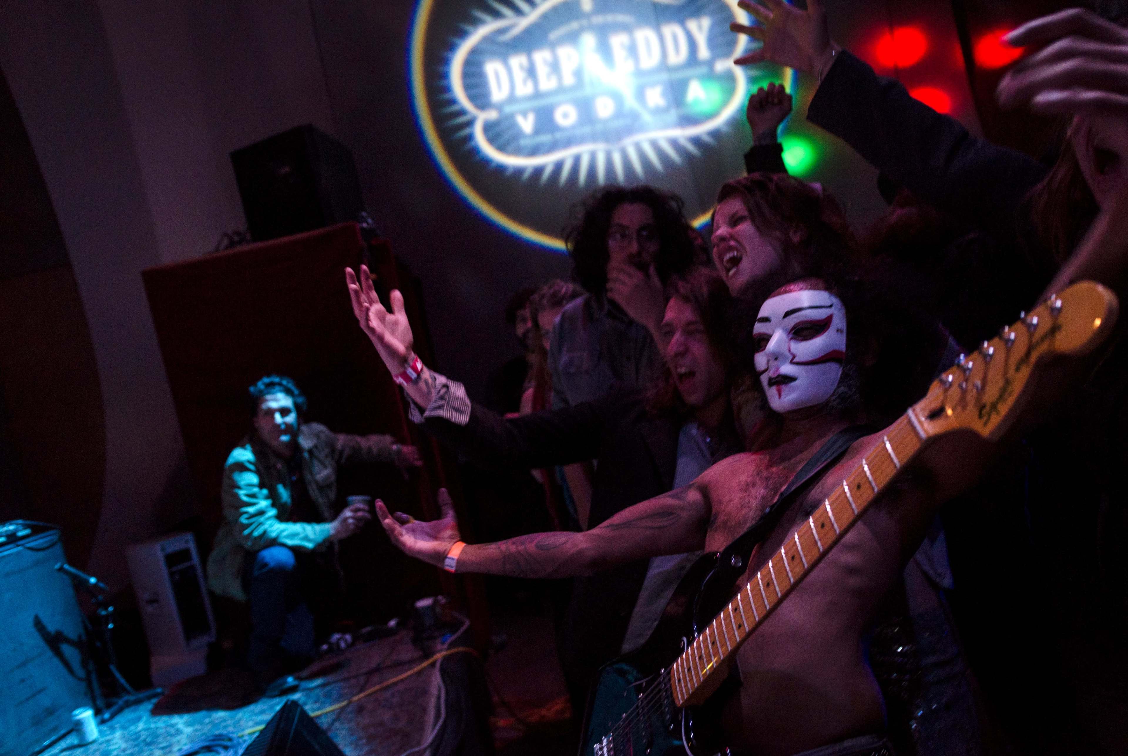 A crowd enjoys a lively performance at a music venue, with a guitarist in face paint and a projection of "Deep Eddy Vodka" on the wall.