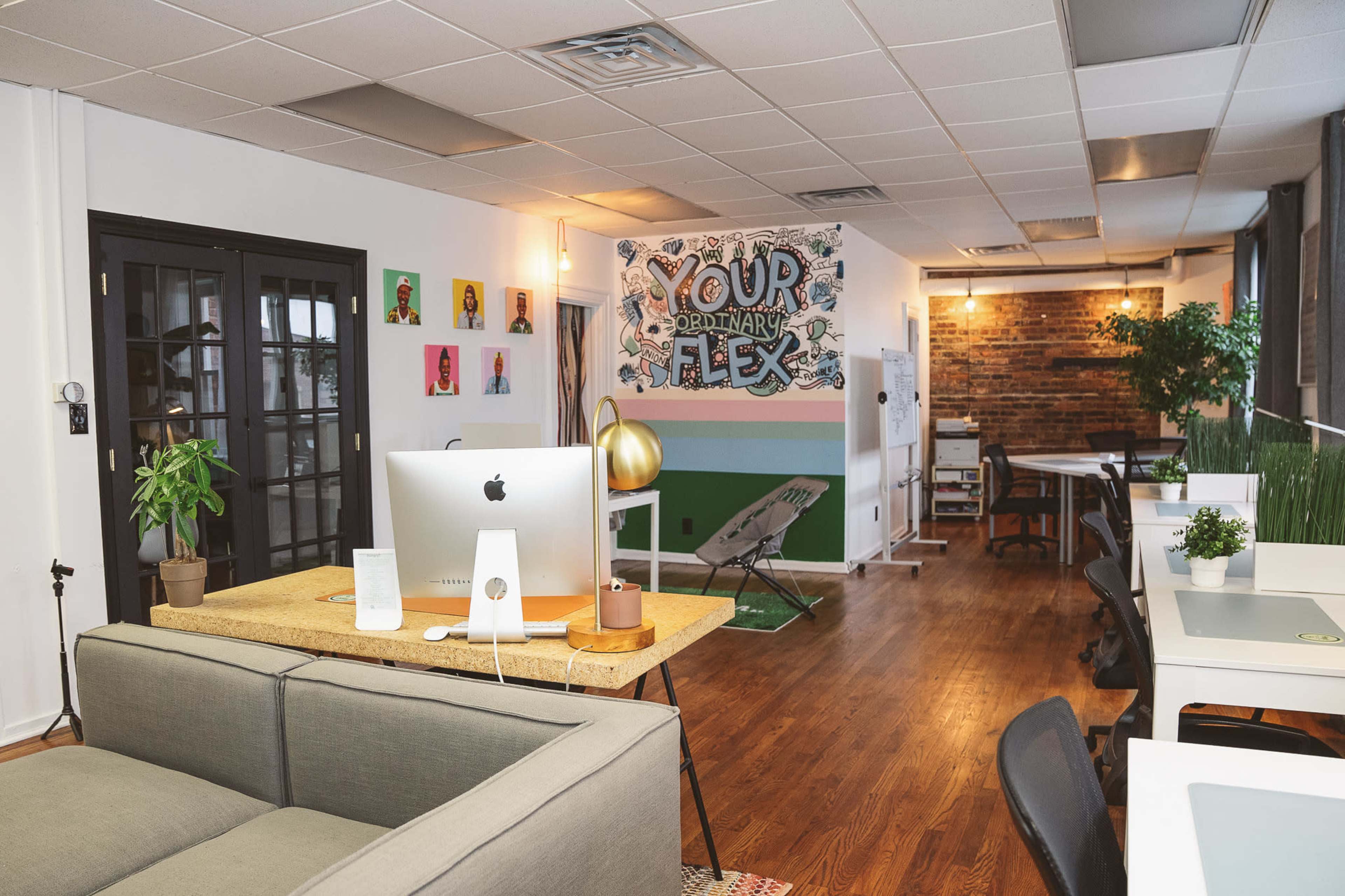 The image shows a modern office space featuring a desk with an iMac, a lounge area with a sofa, colorful wall art, and plants throughout the room.