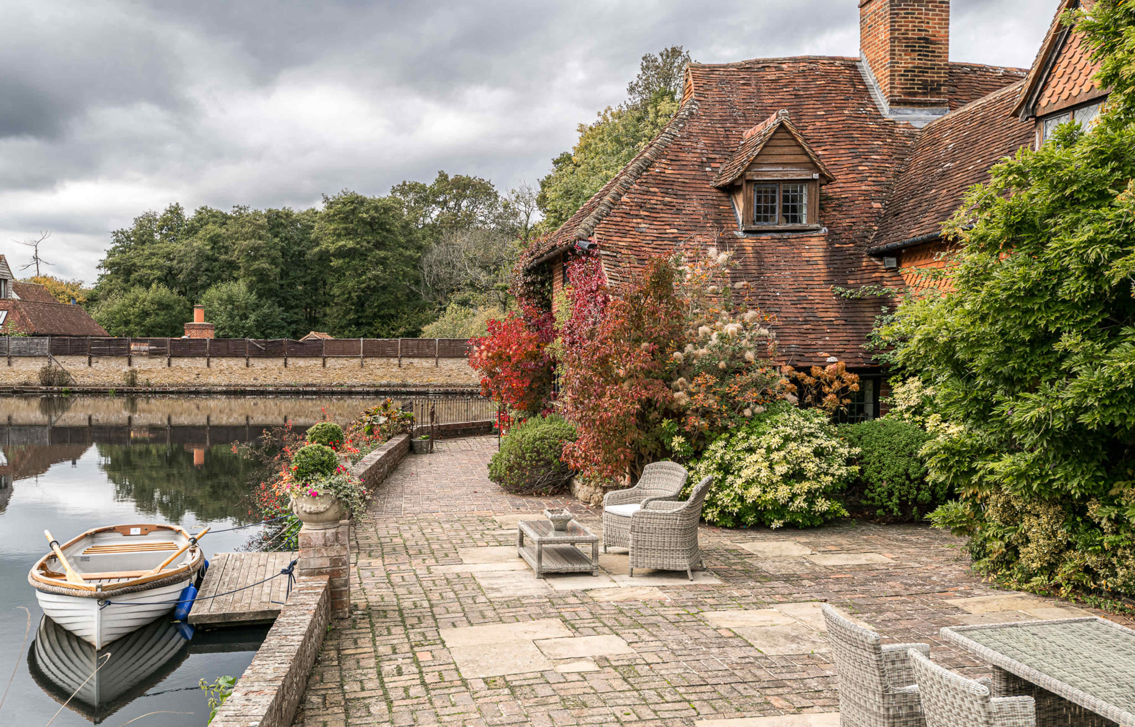 A wooden boat is moored by a stone pathway leading to a brick house, surrounded by greenery and flowers beside a calm body of water.