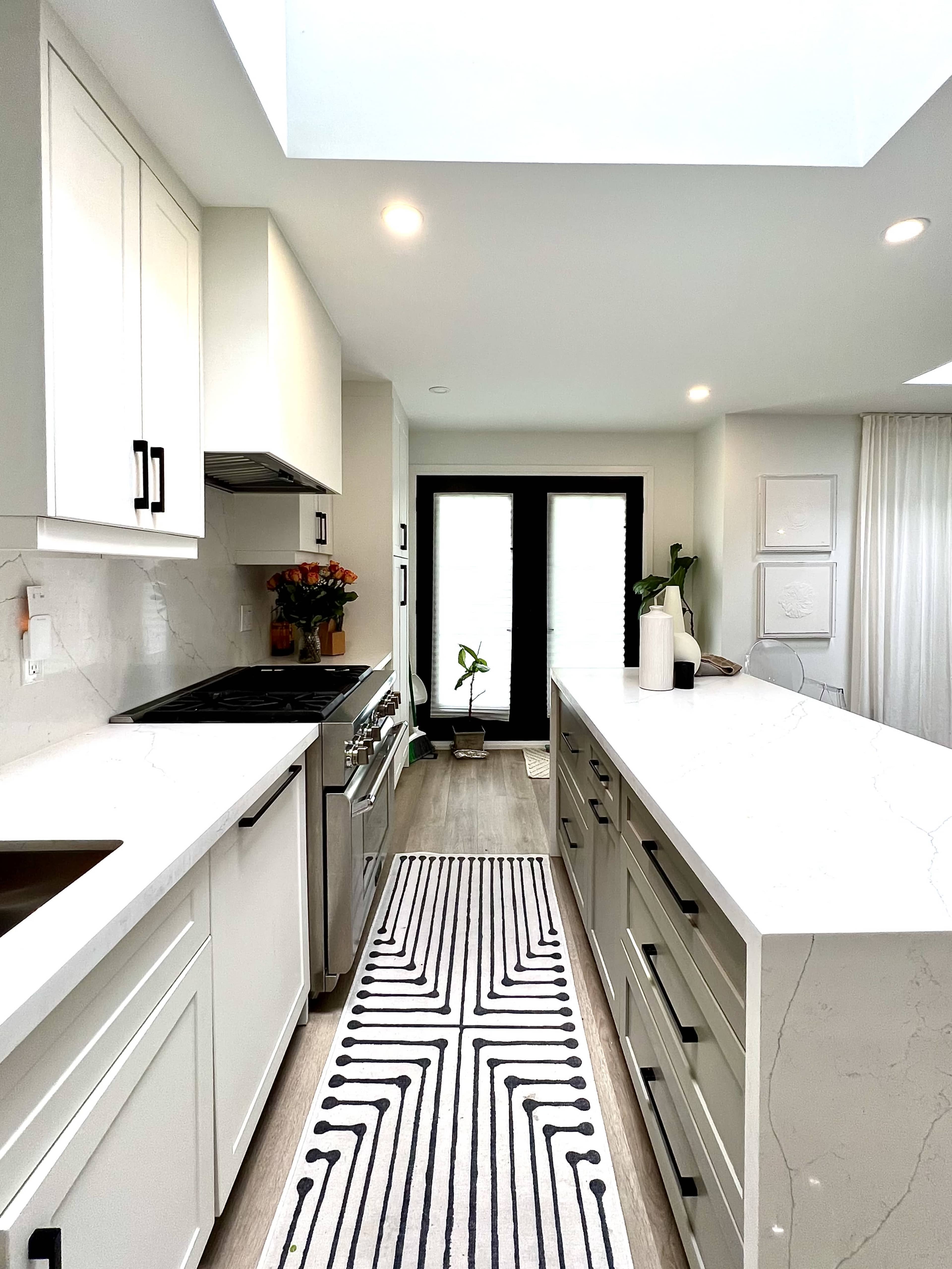 The image shows a modern kitchen with white cabinets, a gray island, a patterned runner rug, and large windows allowing natural light.