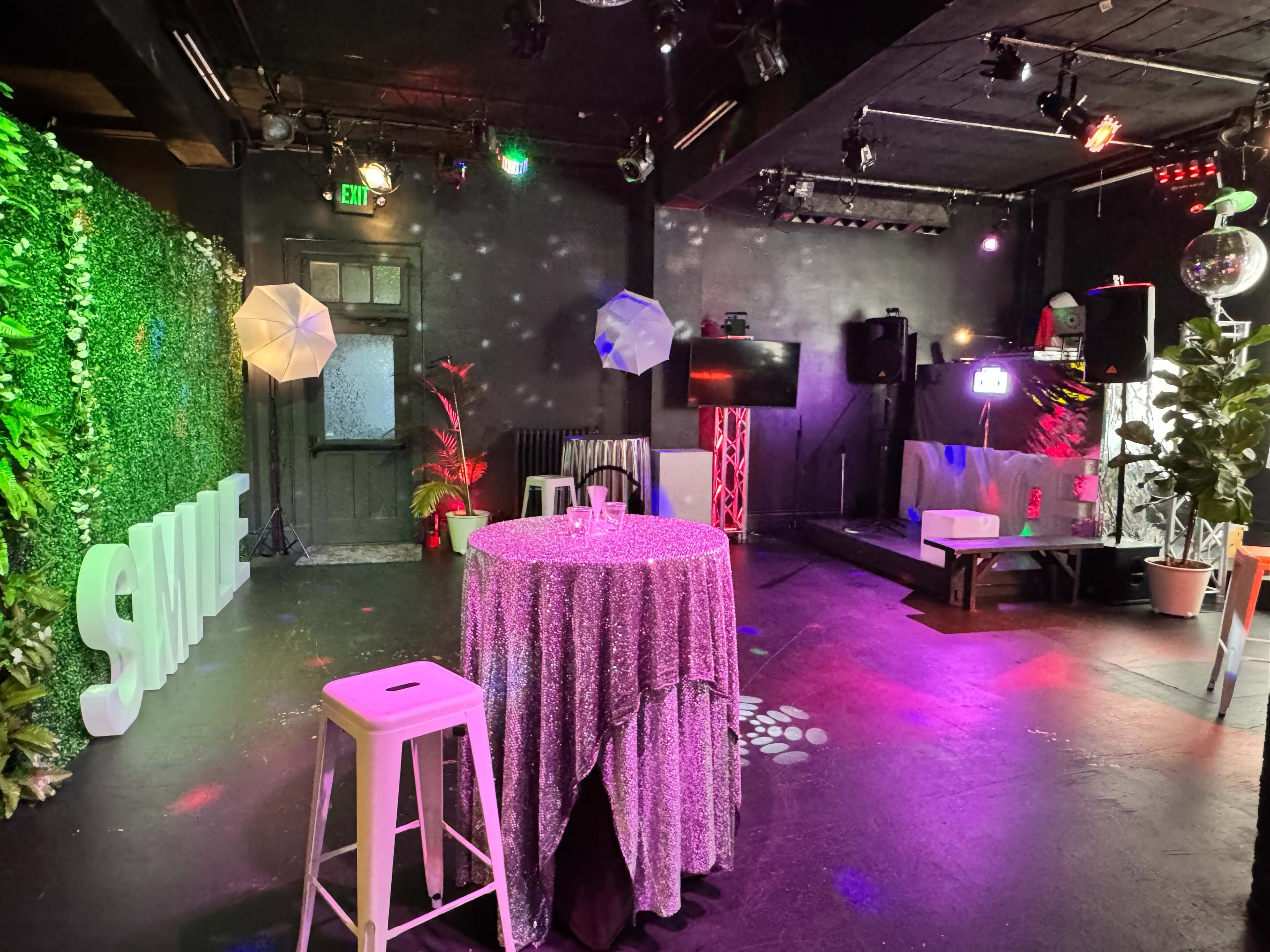 The image shows a decorated event space with a shiny tablecloth, illuminated by colorful lights, featuring plants and various lighting equipment.
