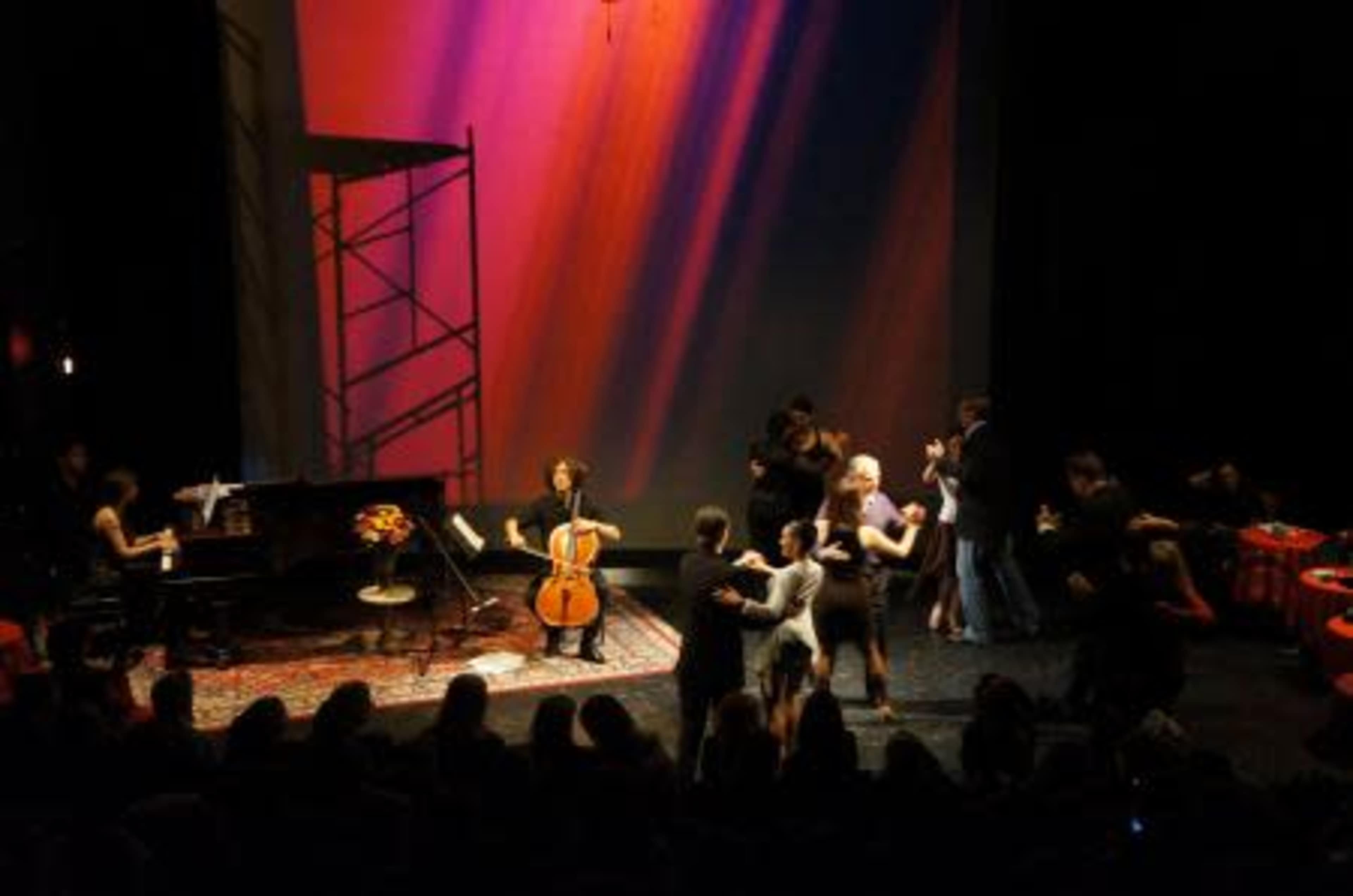 A stage is set with a piano and cello, while people dance in pairs against a colorful backdrop.