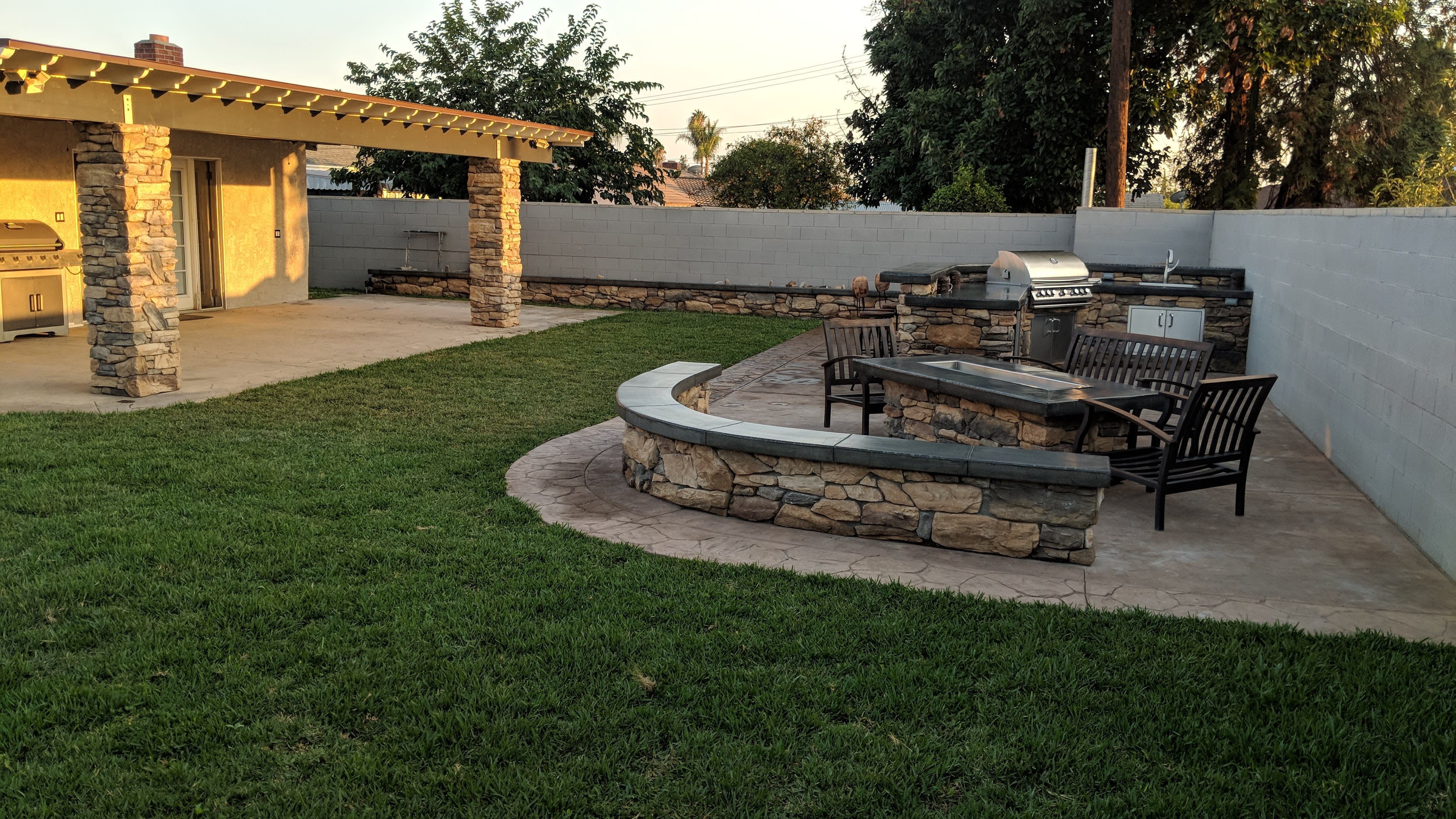 A backyard with a stone patio featuring a circular seating area and a grill setup beside a grassy lawn.