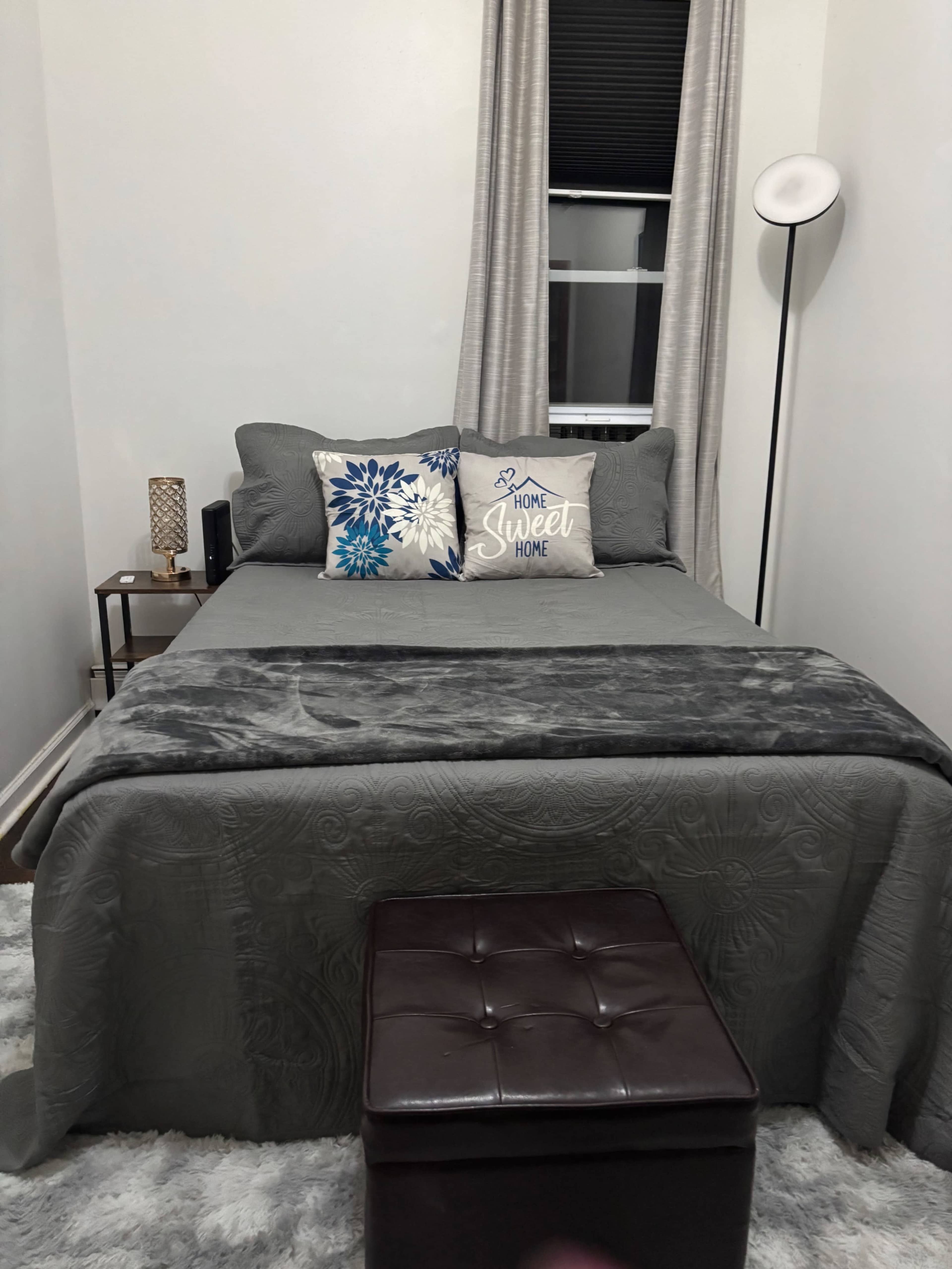 The image shows a neatly made bed in a simple bedroom, featuring gray bedding, decorative pillows, and a small bedside table with a lamp.