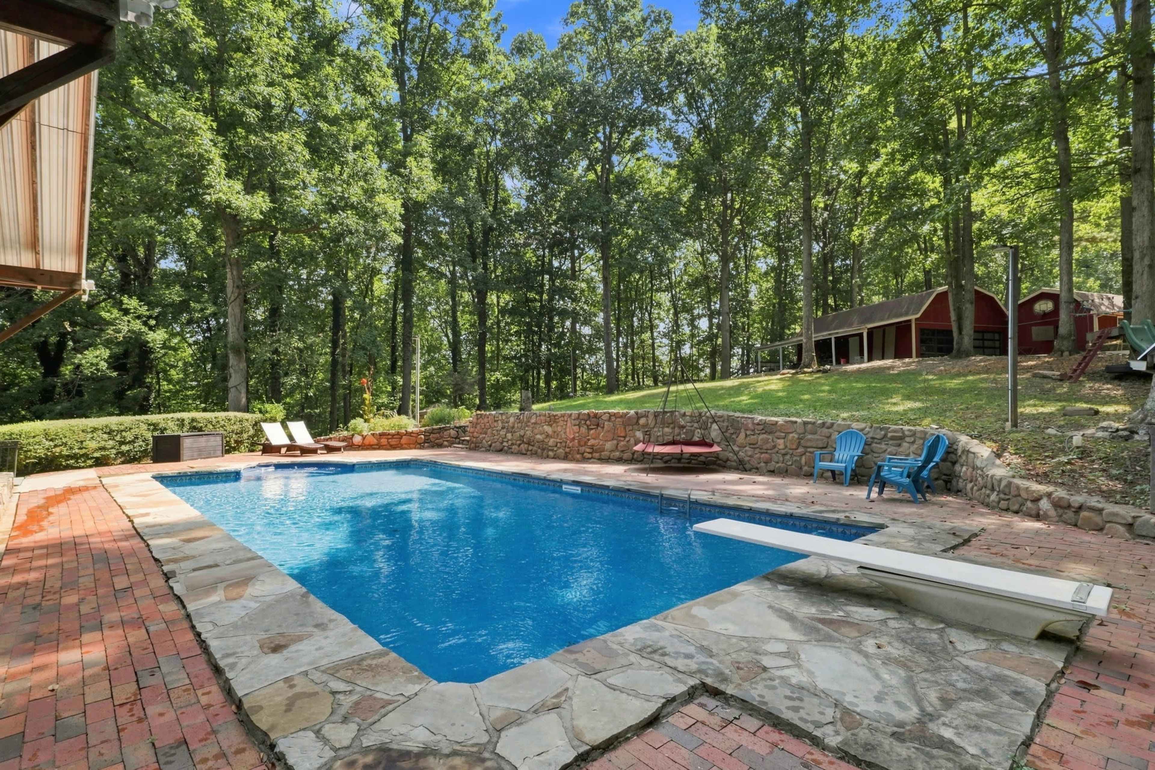 The image shows a rectangular swimming pool surrounded by a stone patio, with blue lounge chairs and trees in the background.