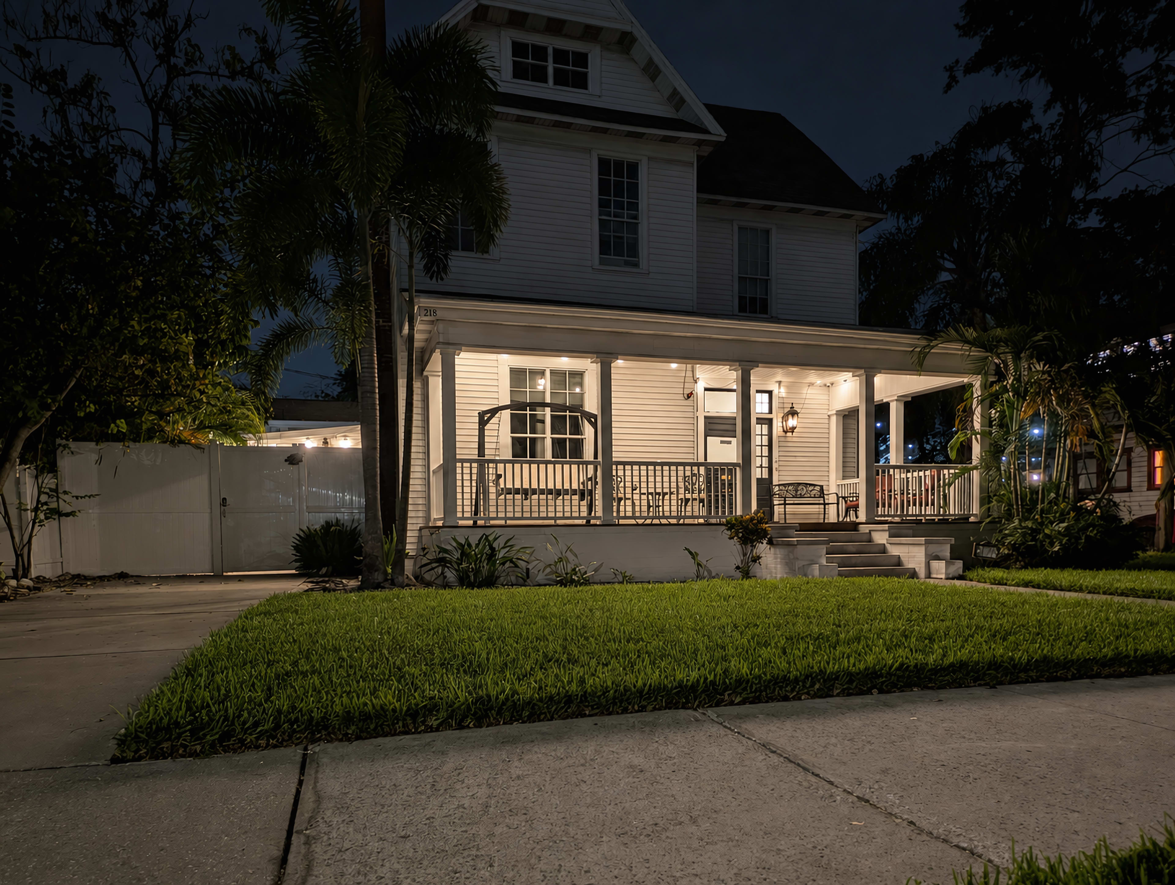 A two-story house with a front porch is illuminated at night, surrounded by well-maintained grass and palm trees.