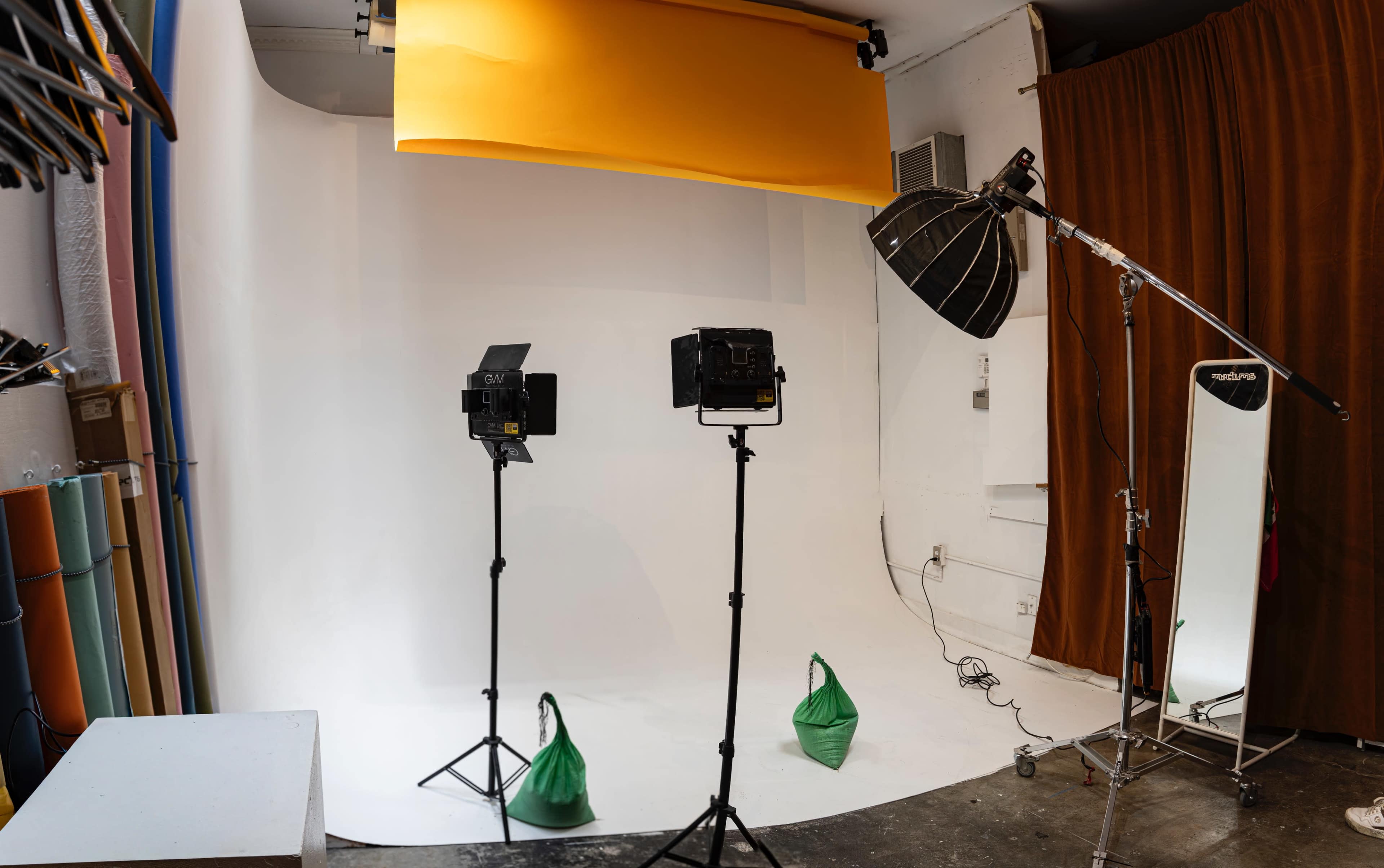 The image shows a photography studio setup with two lights on stands, a white backdrop, and green bags on the floor.