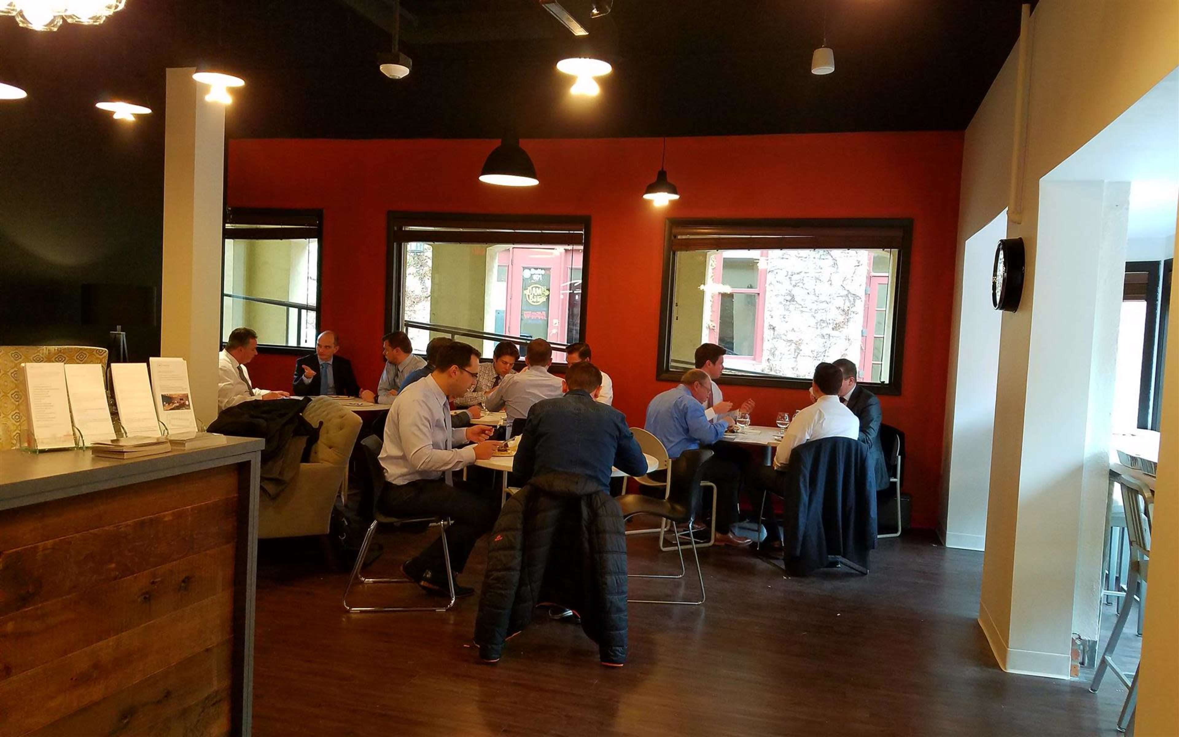 A group of people in business attire sits at tables in a restaurant with large windows and a mix of dark and light decor.