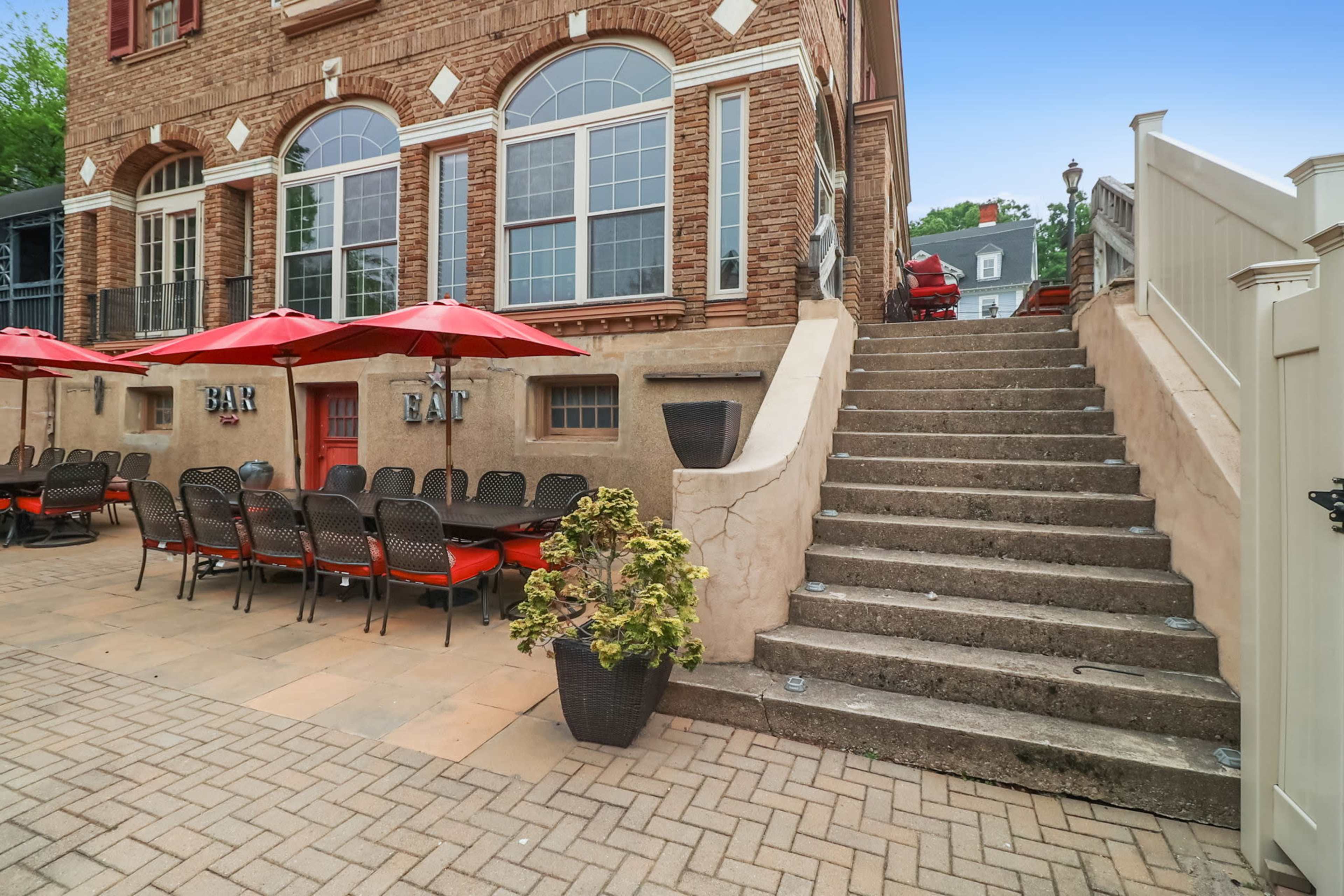 A patio with red umbrellas and outdoor seating leads up to a stairway beside a brick building.
