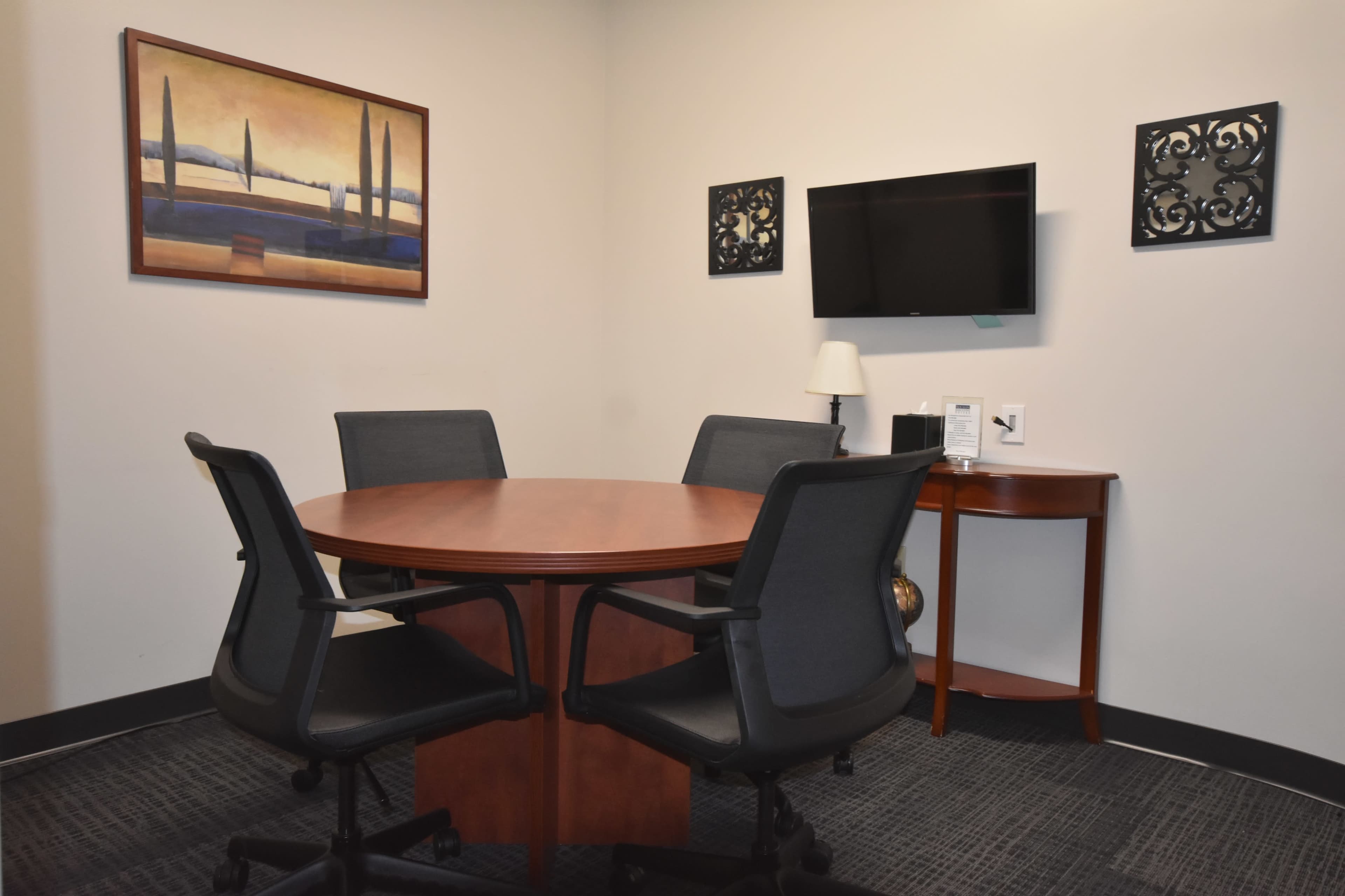 A small conference room features a circular wooden table surrounded by four black chairs, with wall artwork and a mounted television.