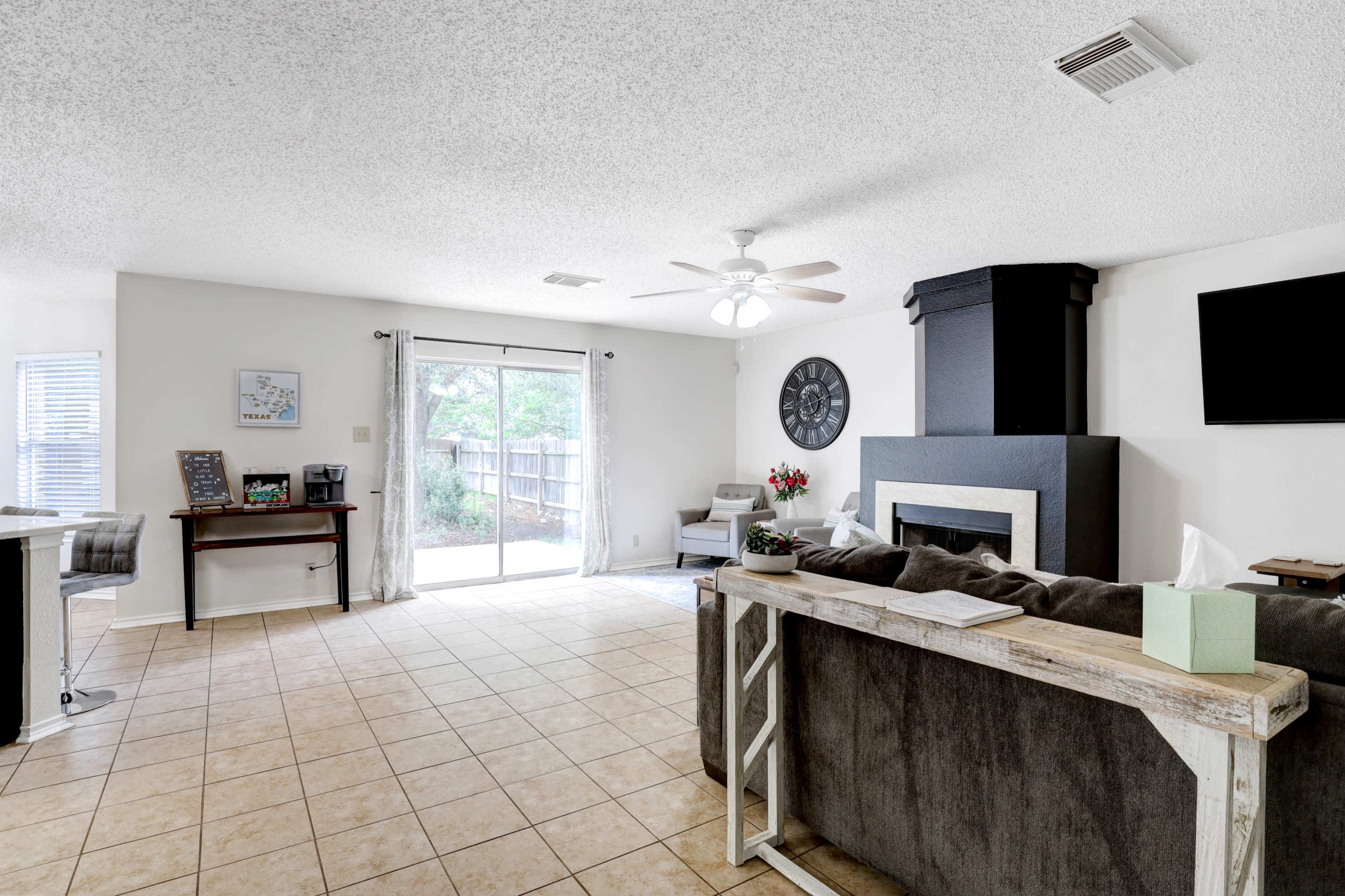 A spacious, well-lit living area with a gray couch, a flat-screen TV on the wall, a fireplace, and large windows leading to an outdoor space.