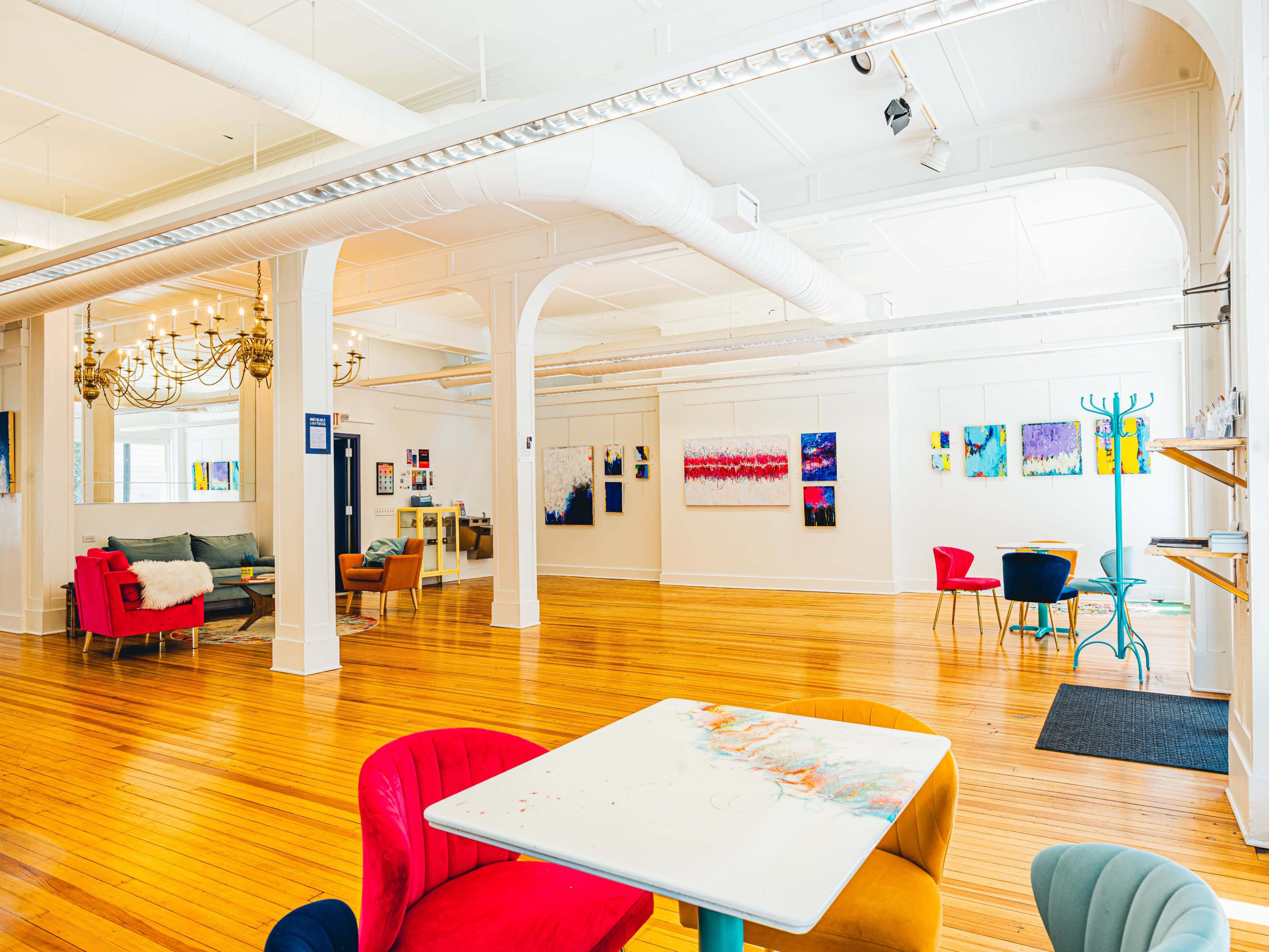The image shows a spacious art studio with wooden floors, white walls adorned with colorful paintings, and a mix of modern furniture, including red and blue chairs and a table.