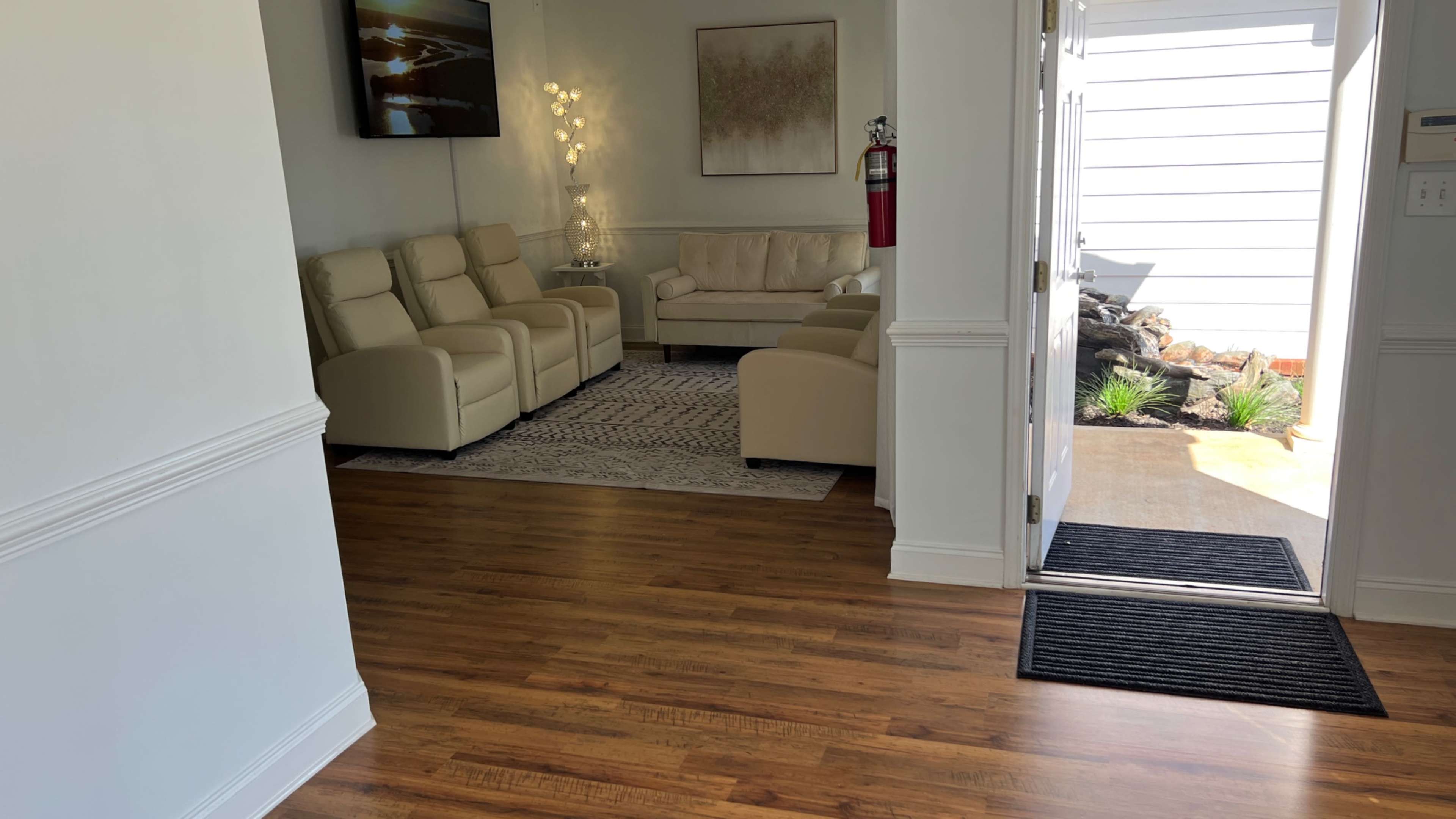 The image shows a bright, modern living area with light-colored furniture and a wooden floor, leading to an entrance that opens to an outdoor pathway.