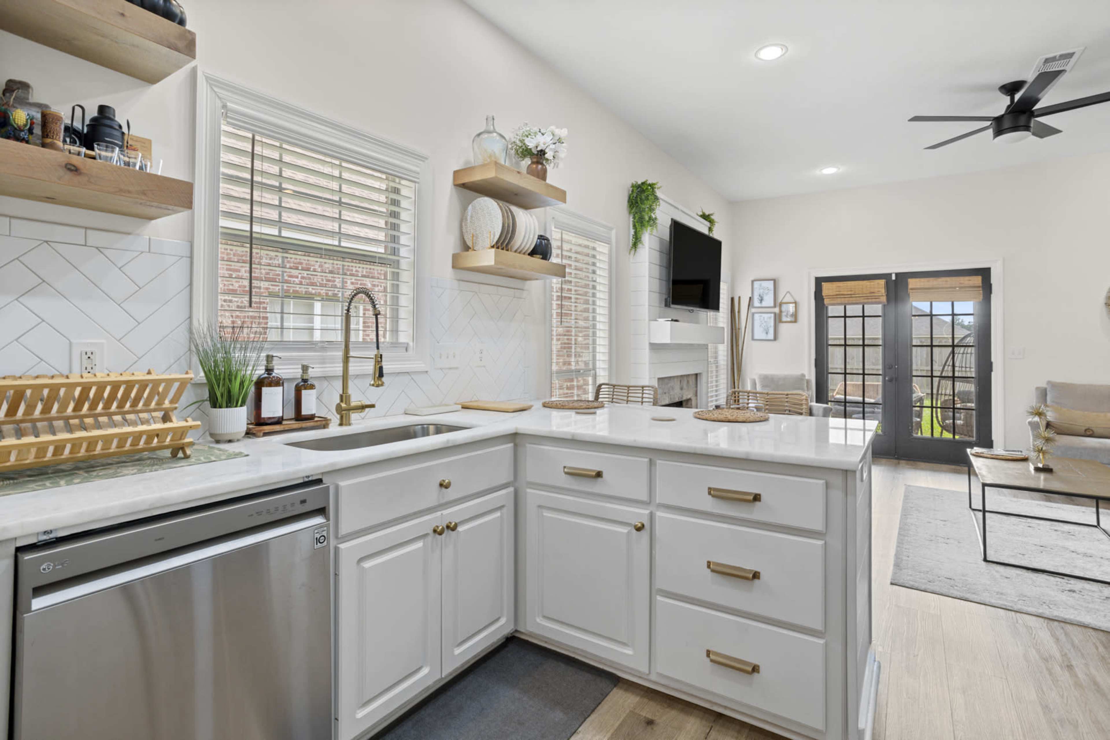 The image shows a modern kitchen featuring white cabinetry, a stainless steel dishwasher, a light countertop, and open shelving with decorative items.