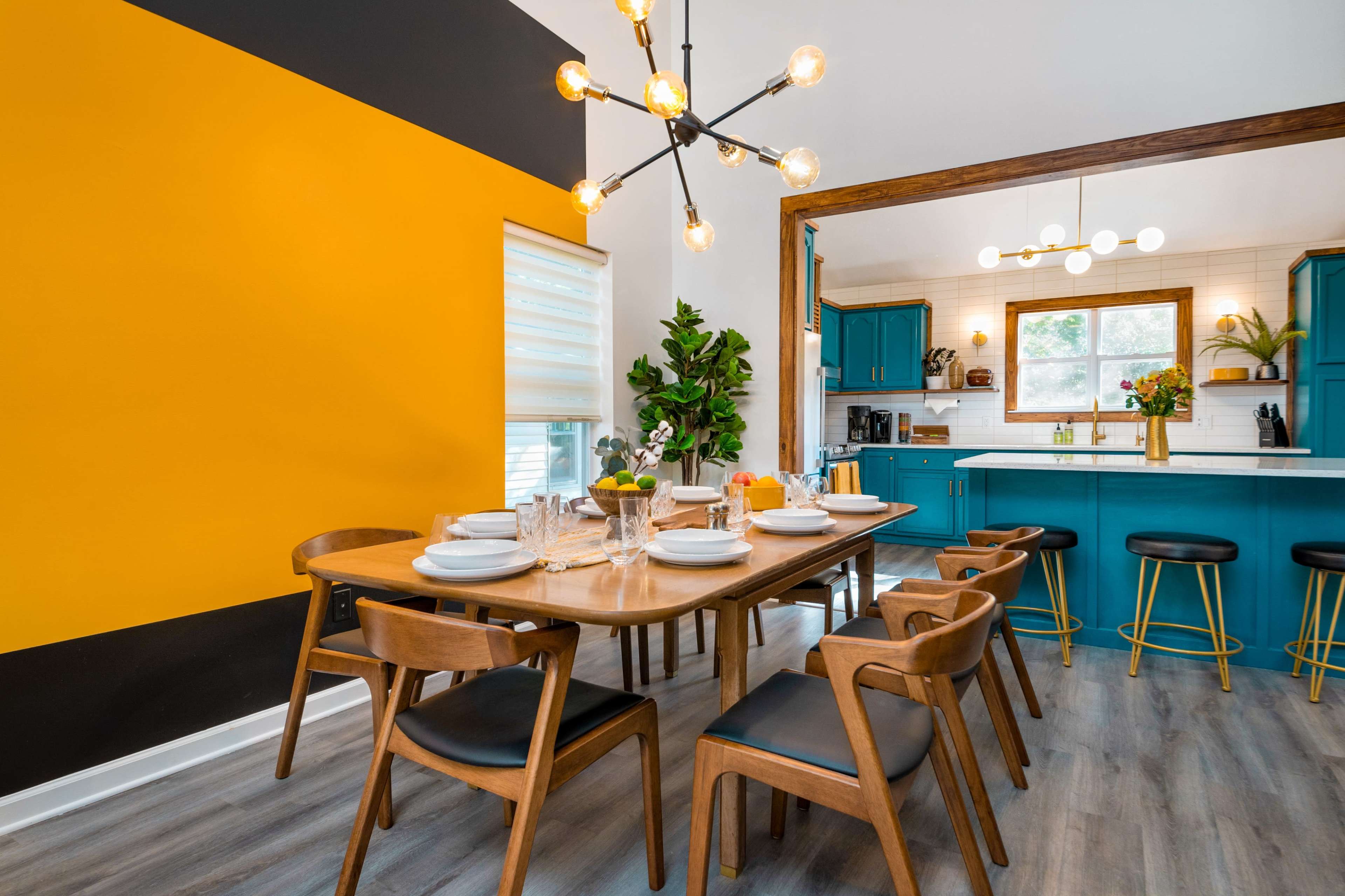 The dining area features a wooden table set with plates and glasses, surrounded by chairs, and is complemented by a bright yellow accent wall and teal cabinetry in the adjacent kitchen.