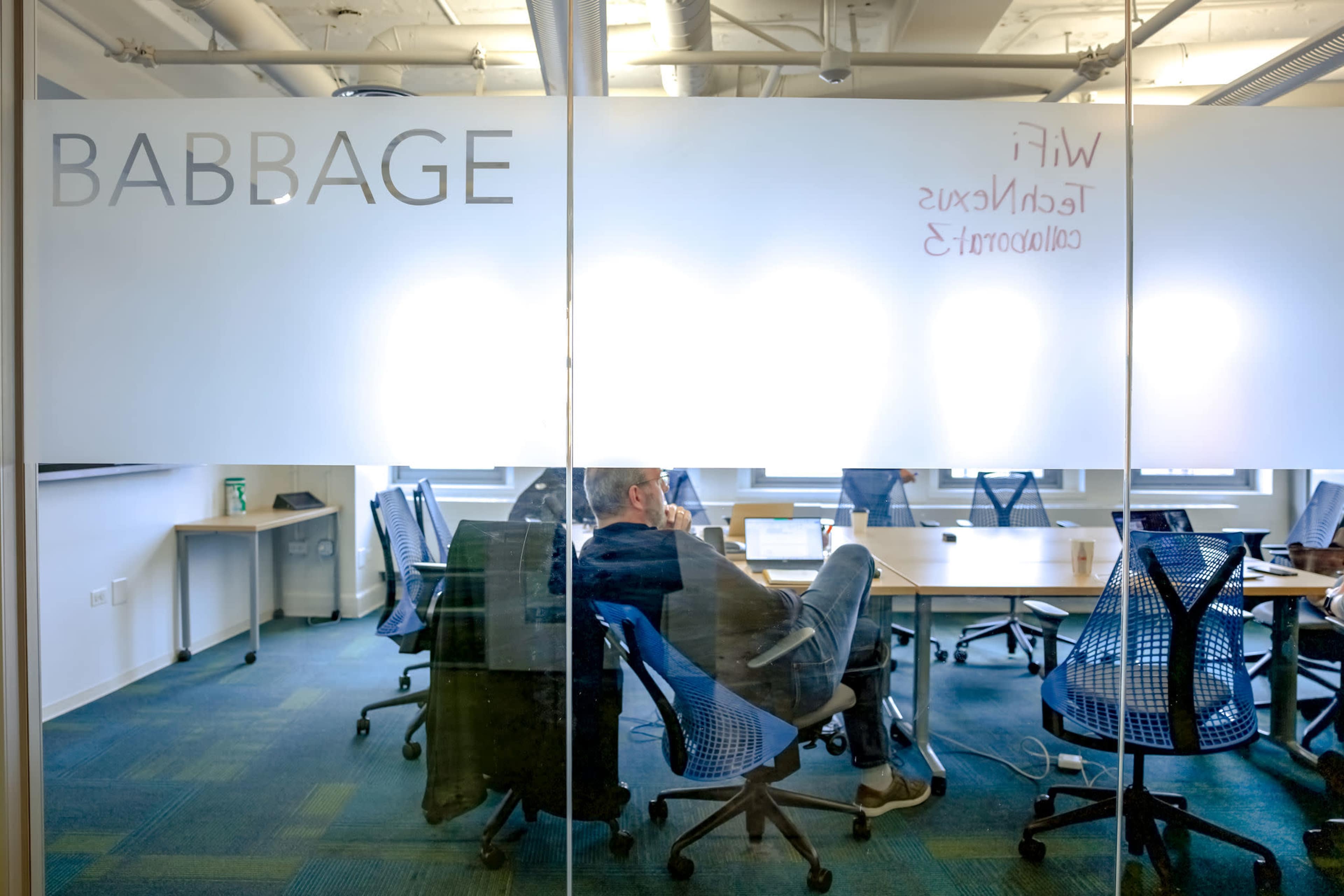 A person sits in a chair with their feet on a table in a glass-walled office labeled "BABBAGE," with scattered chairs and a whiteboard visible in the background.