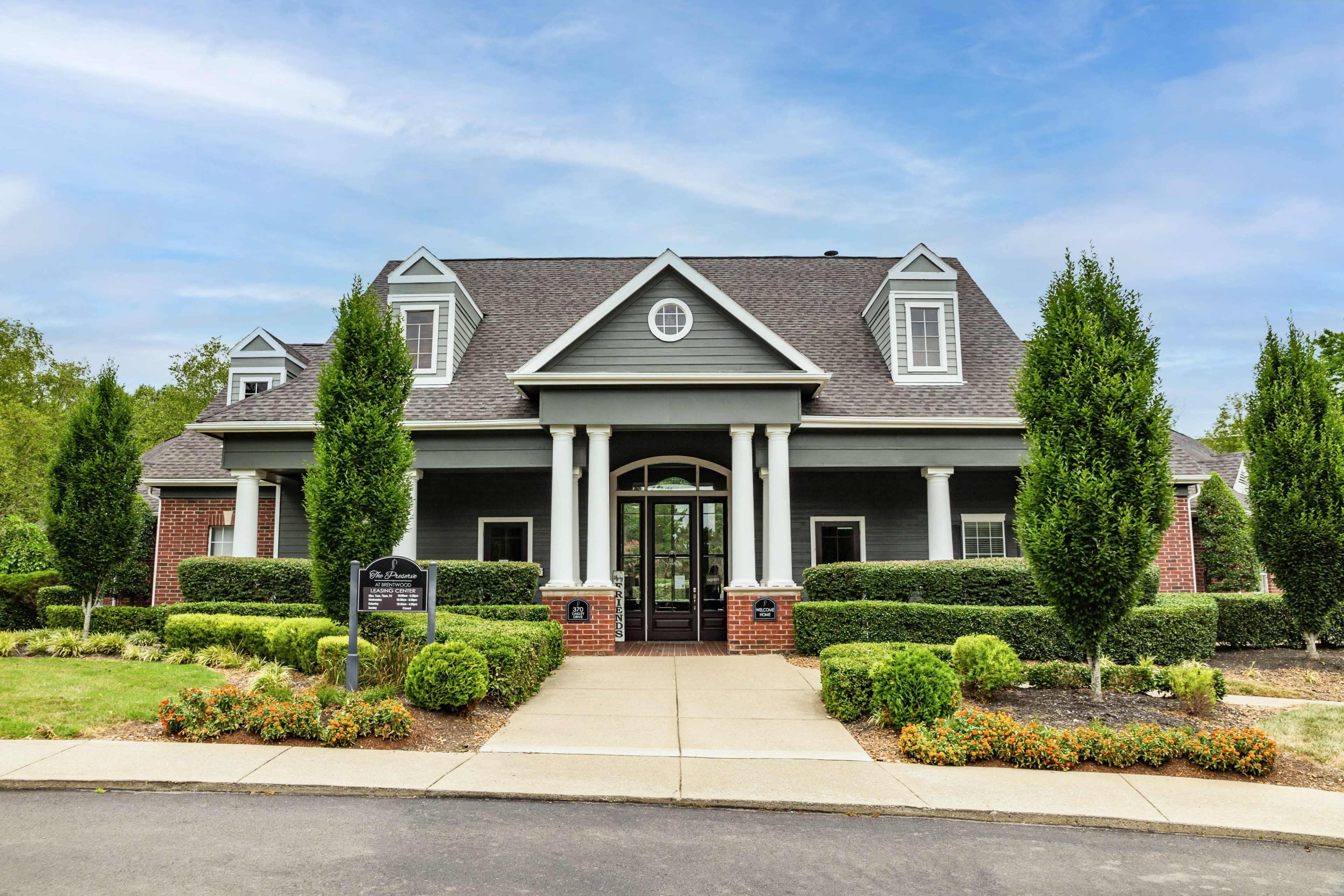 The image shows a large gray house with a gabled roof, flanked by well-manicured shrubs and trees, situated on a paved driveway.