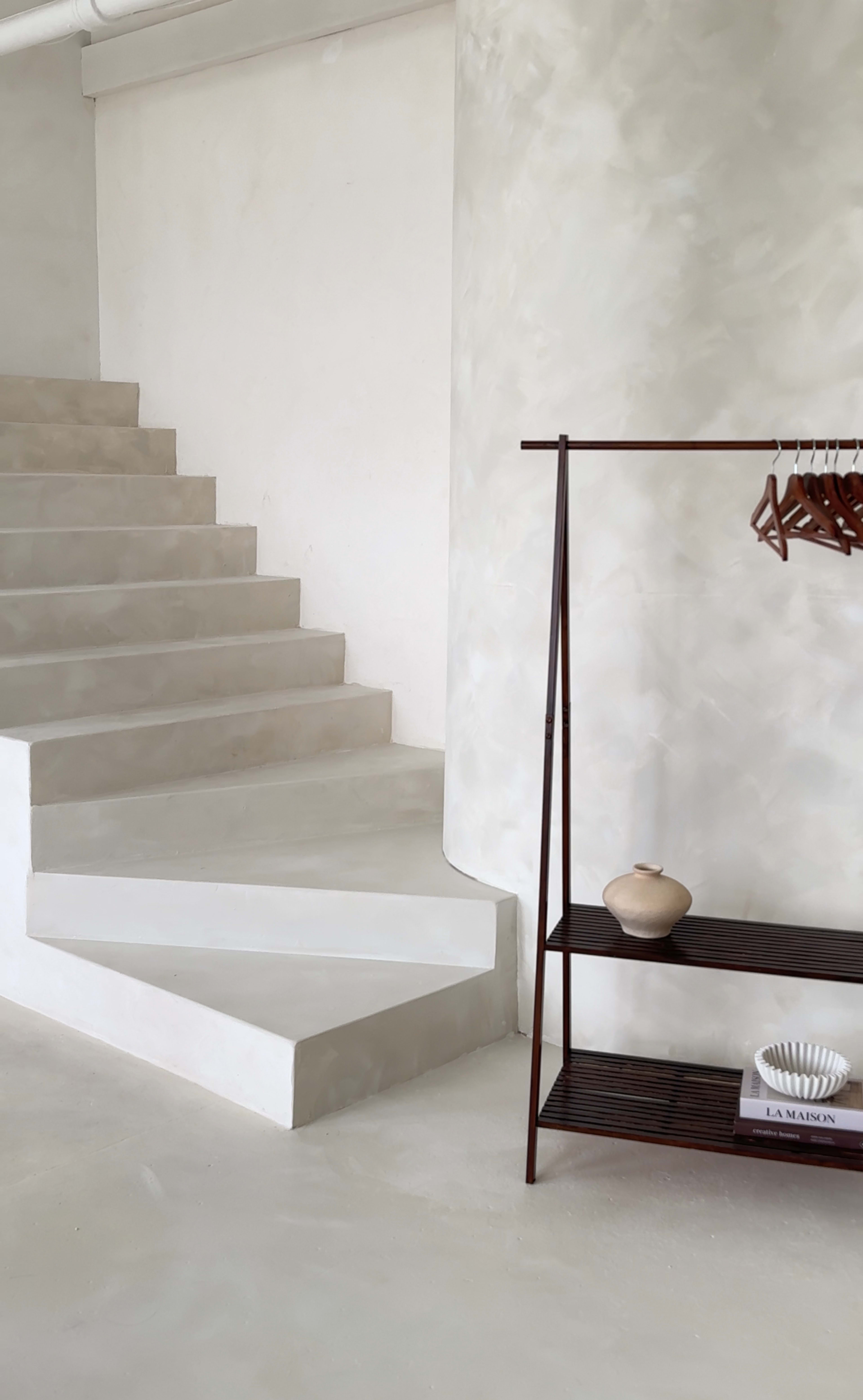 A minimalist interior features a smooth, curved staircase leading upwards, alongside a sleek coat rack with wooden hangers and a small shelf holding decorative items.