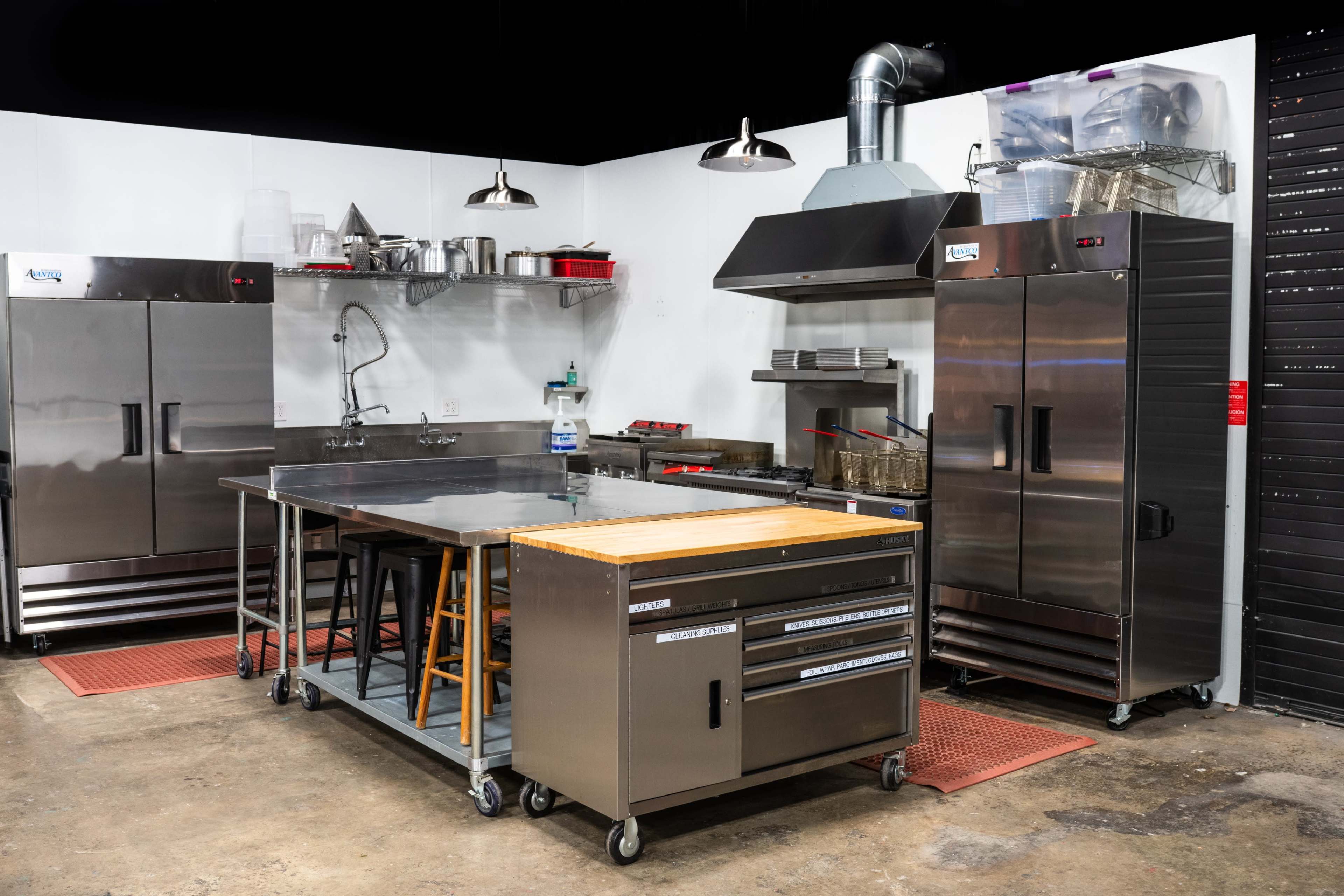 A commercial kitchen equipped with stainless steel appliances, including refrigerators, a stove hood, and a work table with a wooden surface.