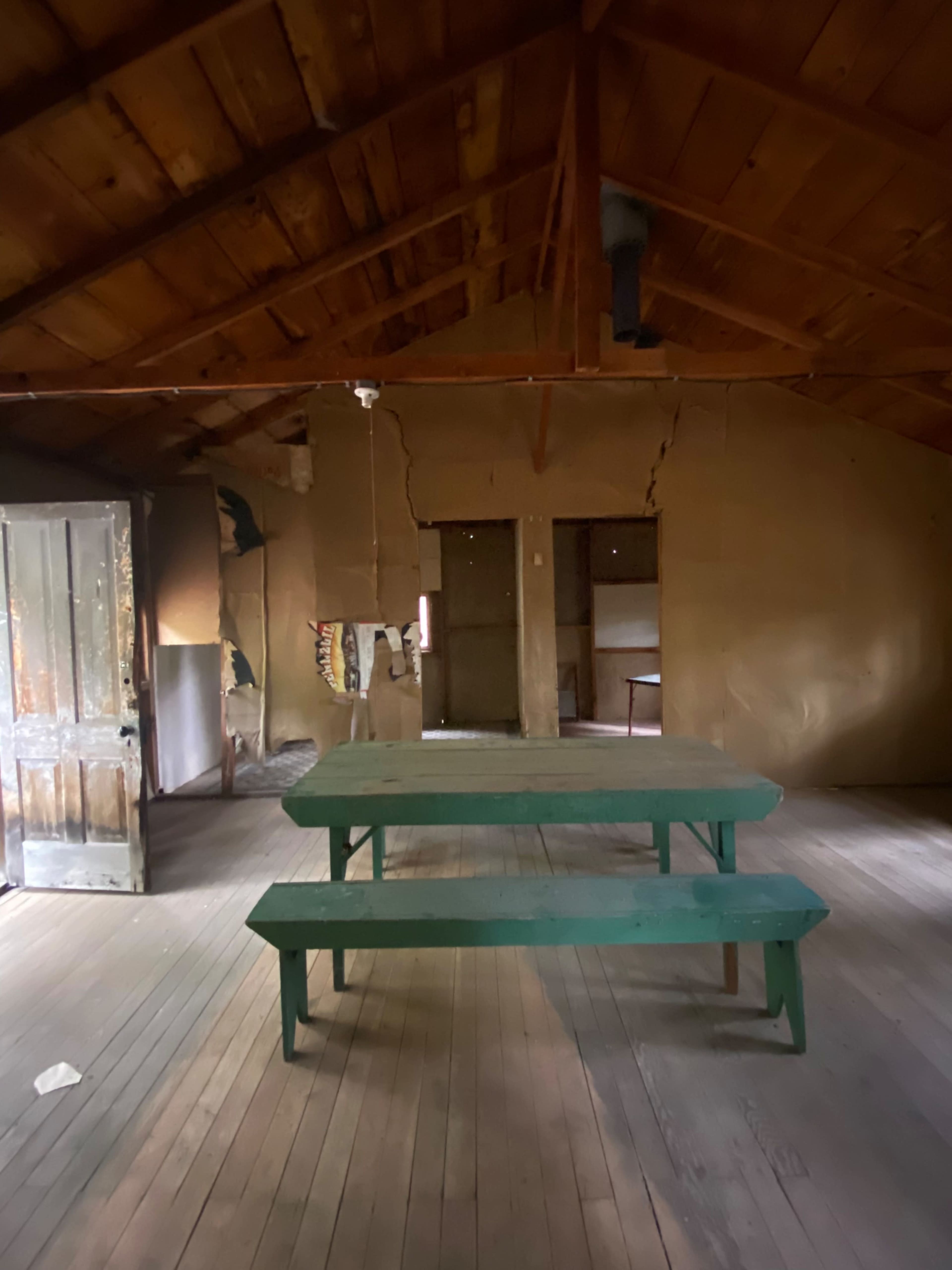 An empty room features a green wooden table with benches beneath a sloped wooden ceiling.