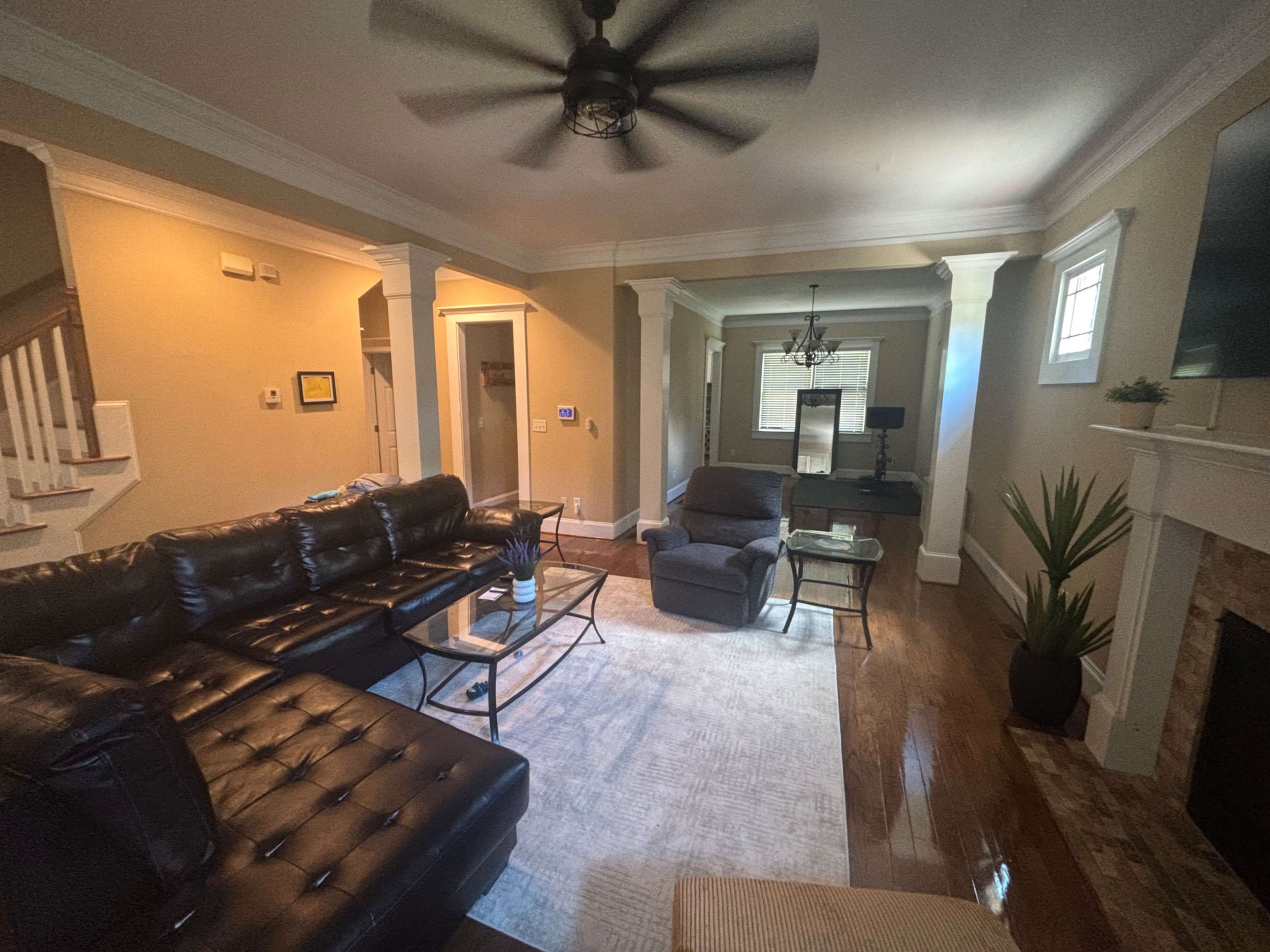 The image shows a spacious living room featuring a leather sectional sofa, a glass coffee table, and a recliner chair, with hardwood floors and light-colored walls.