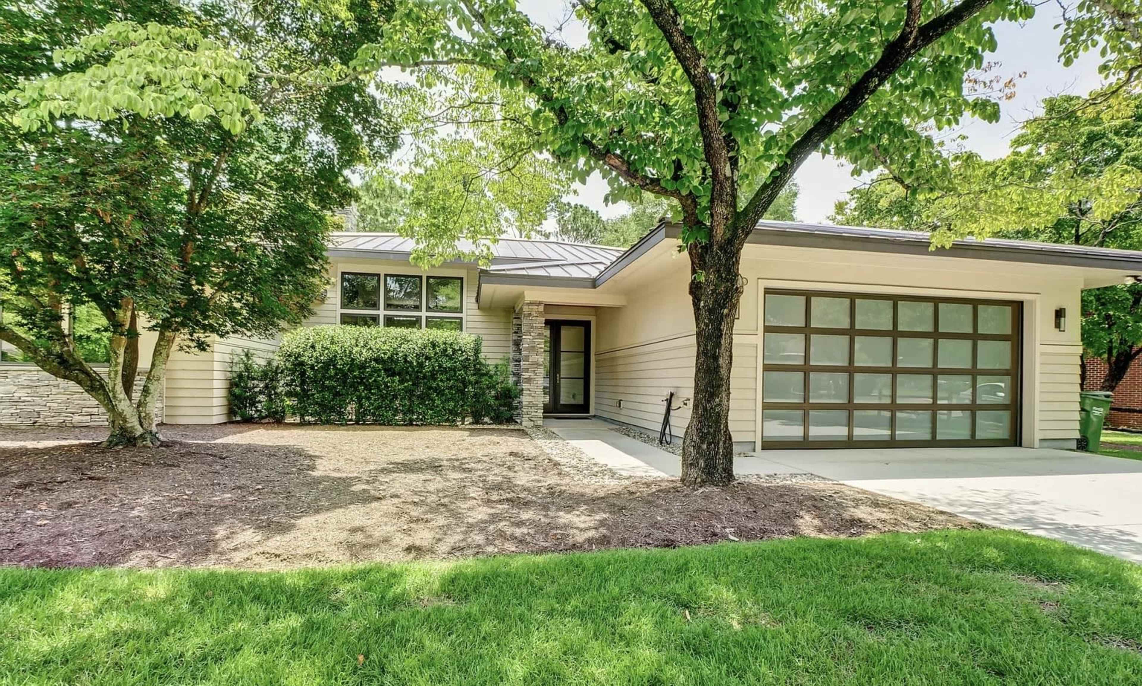 A modern house with a stone accent wall and large windows, set on a grassy lawn and surrounded by trees.