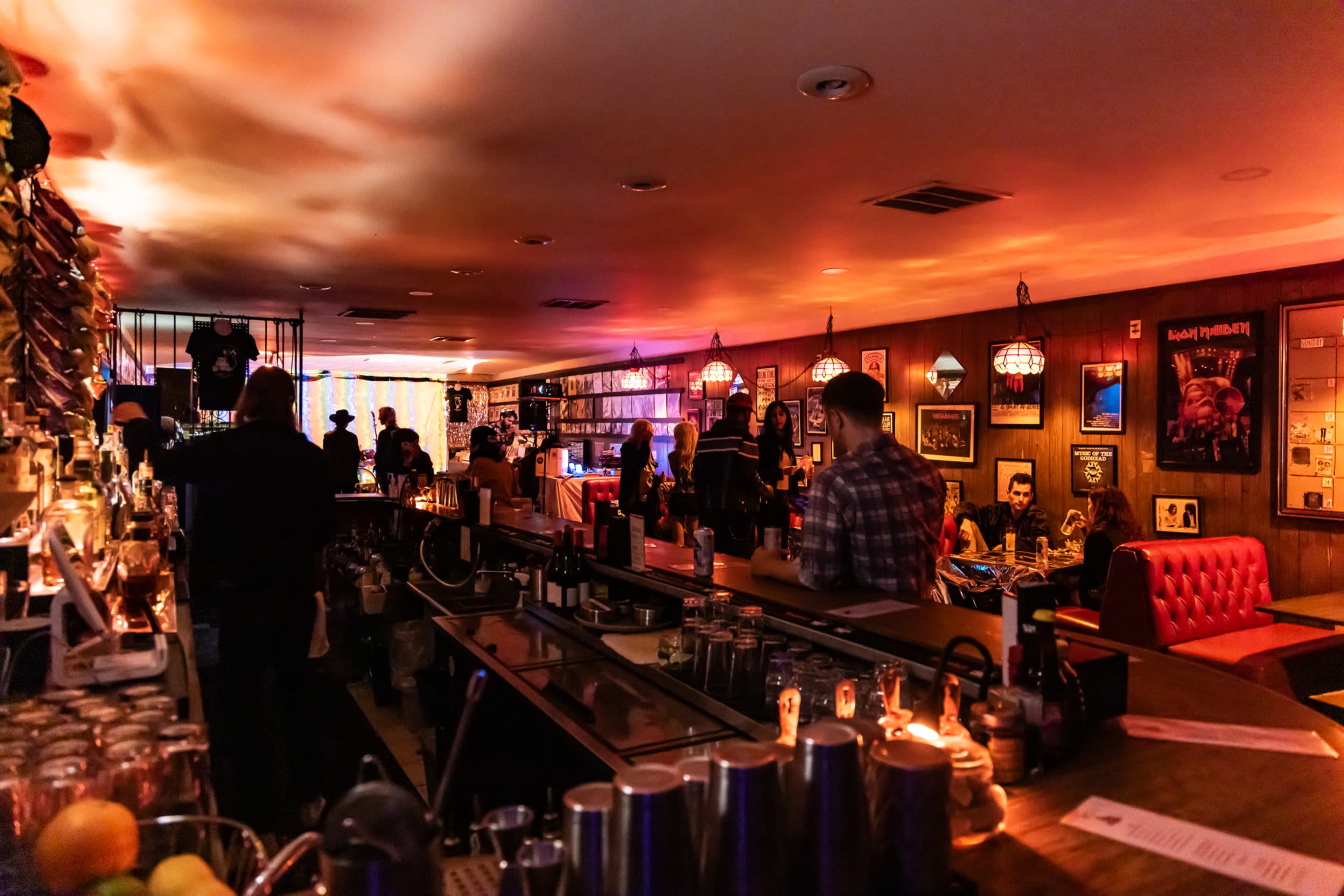 A dimly lit bar interior features patrons mingling, a long bar stocked with drinks, and walls adorned with framed posters and warm lighting fixtures.