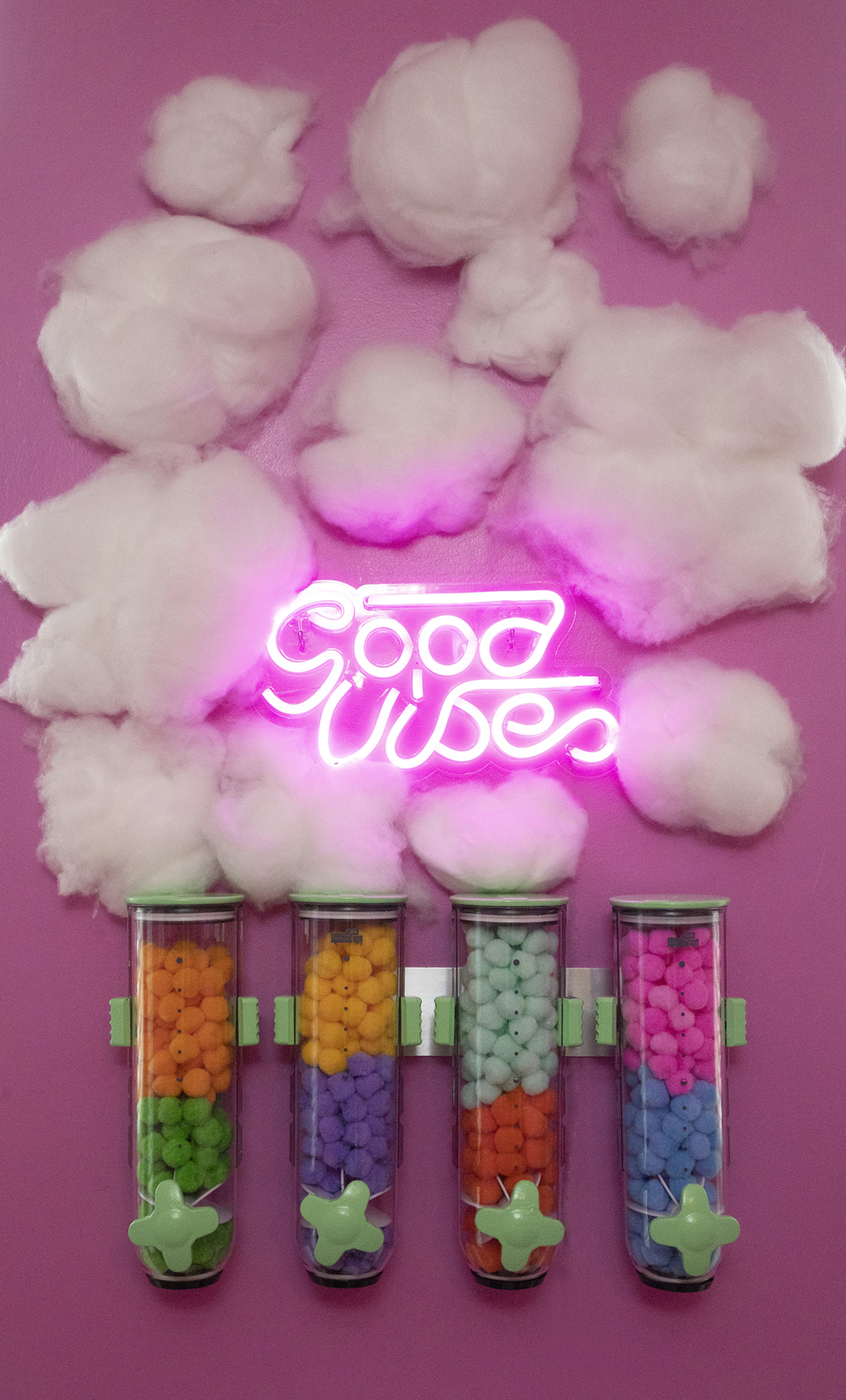 A wall decorated with fluffy white cotton clouds and a neon sign that reads "Good Vibes," above four clear tubes filled with colorful pom-poms.