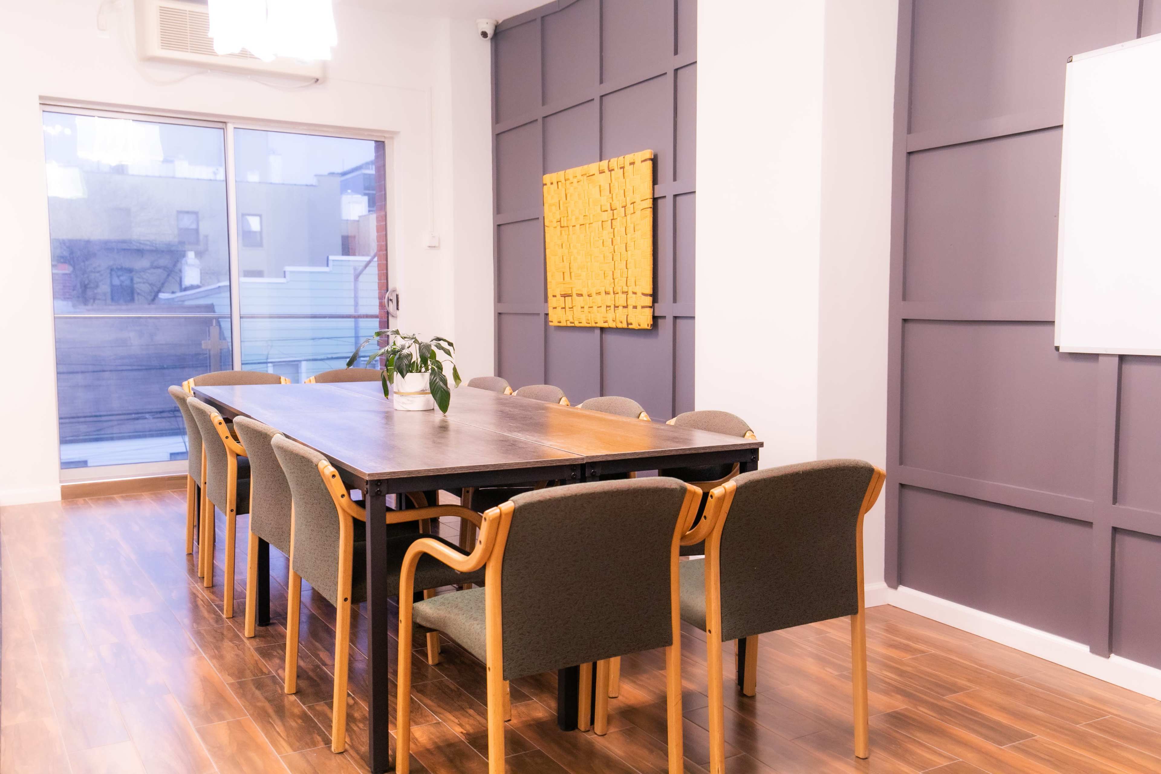 A large conference room features a long wooden table surrounded by ten chairs, with a plant on the table and a large window overlooking an exterior balcony.
