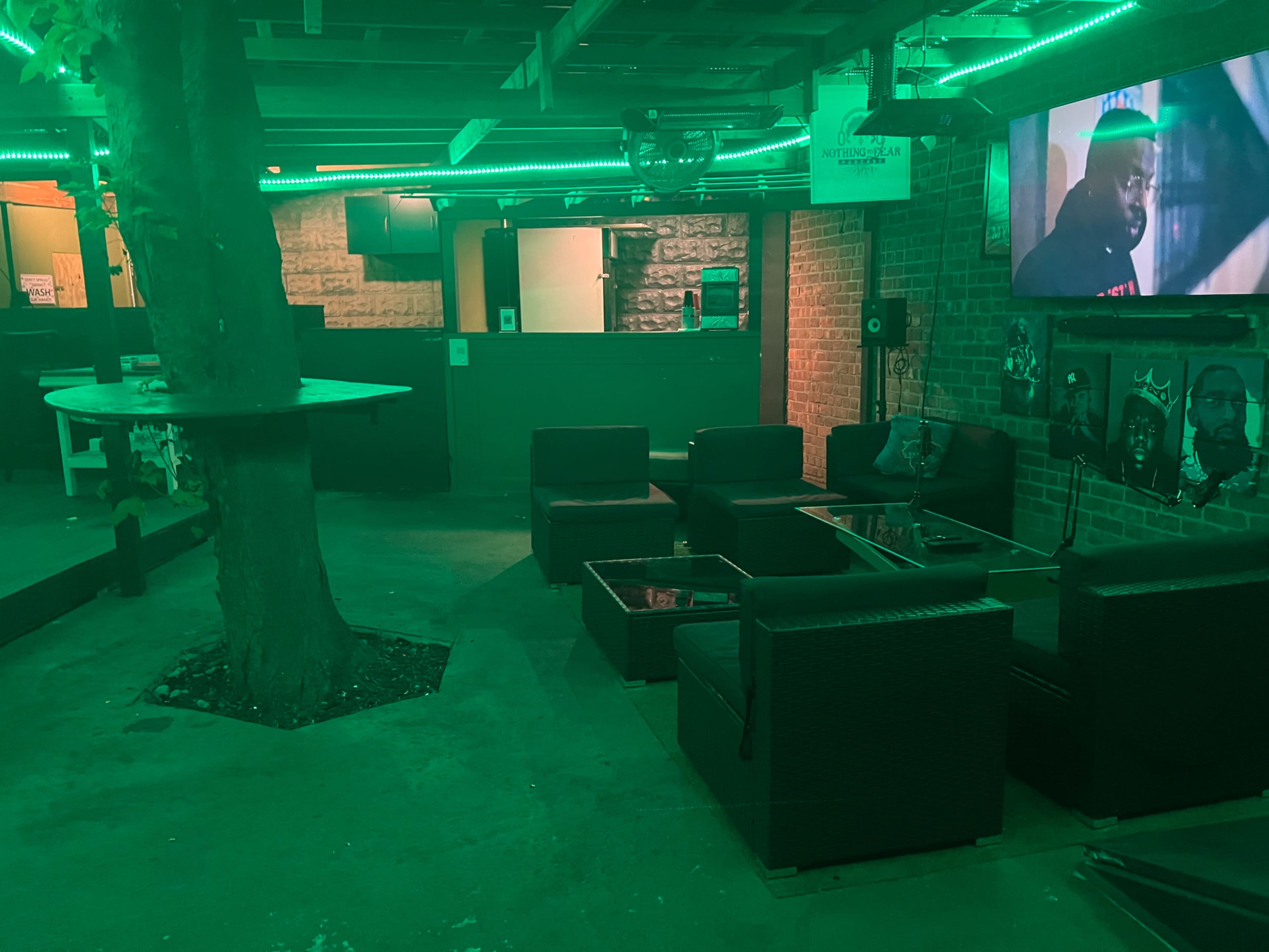 A dimly lit lounge area with green LED lighting, featuring several black wicker chairs and glass tables near a tree, with a large screen displaying a video on the wall.