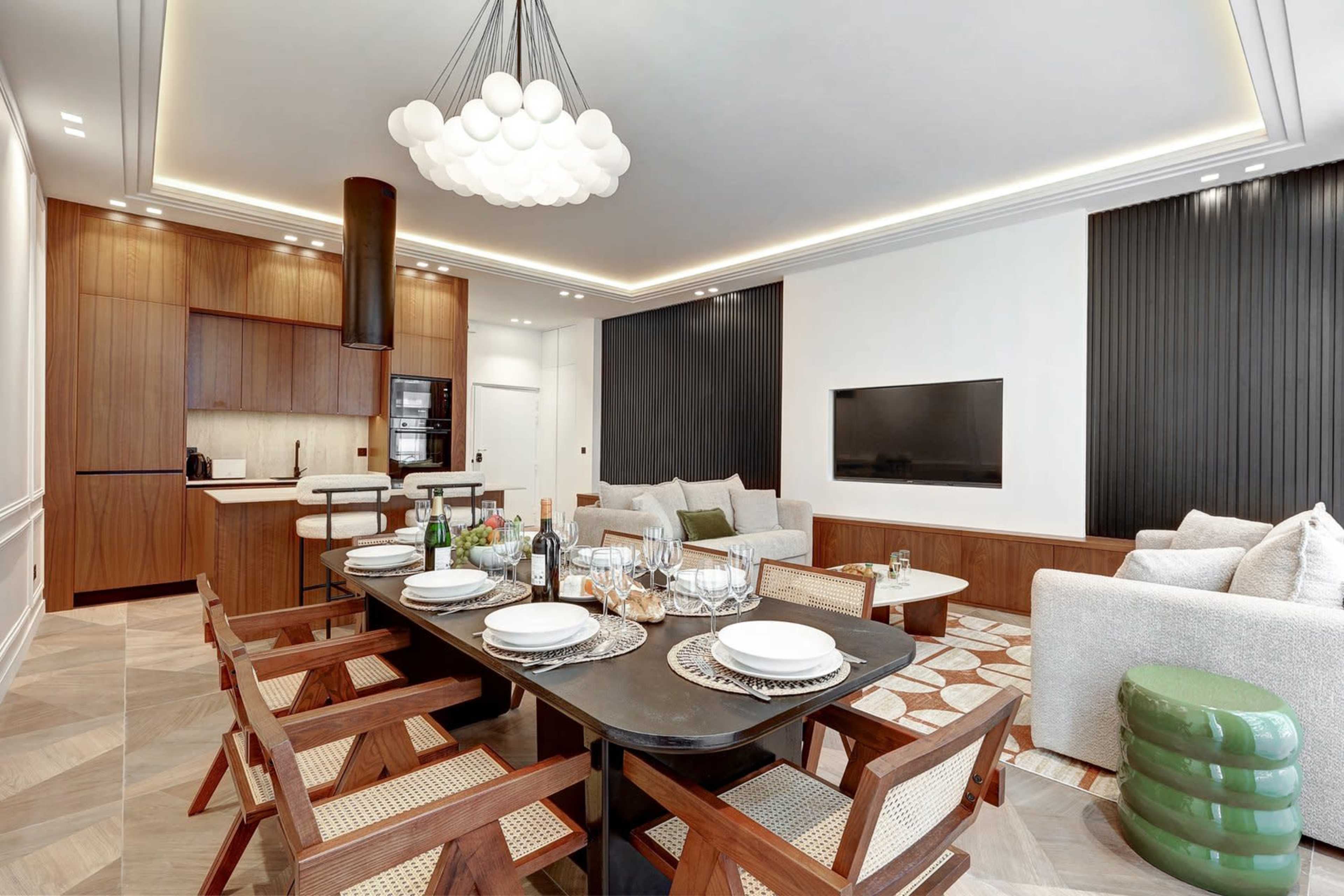 The image shows a modern dining area with a large table set for a meal, surrounded by wooden chairs, and a living space featuring a sofa and television in a contemporary design.