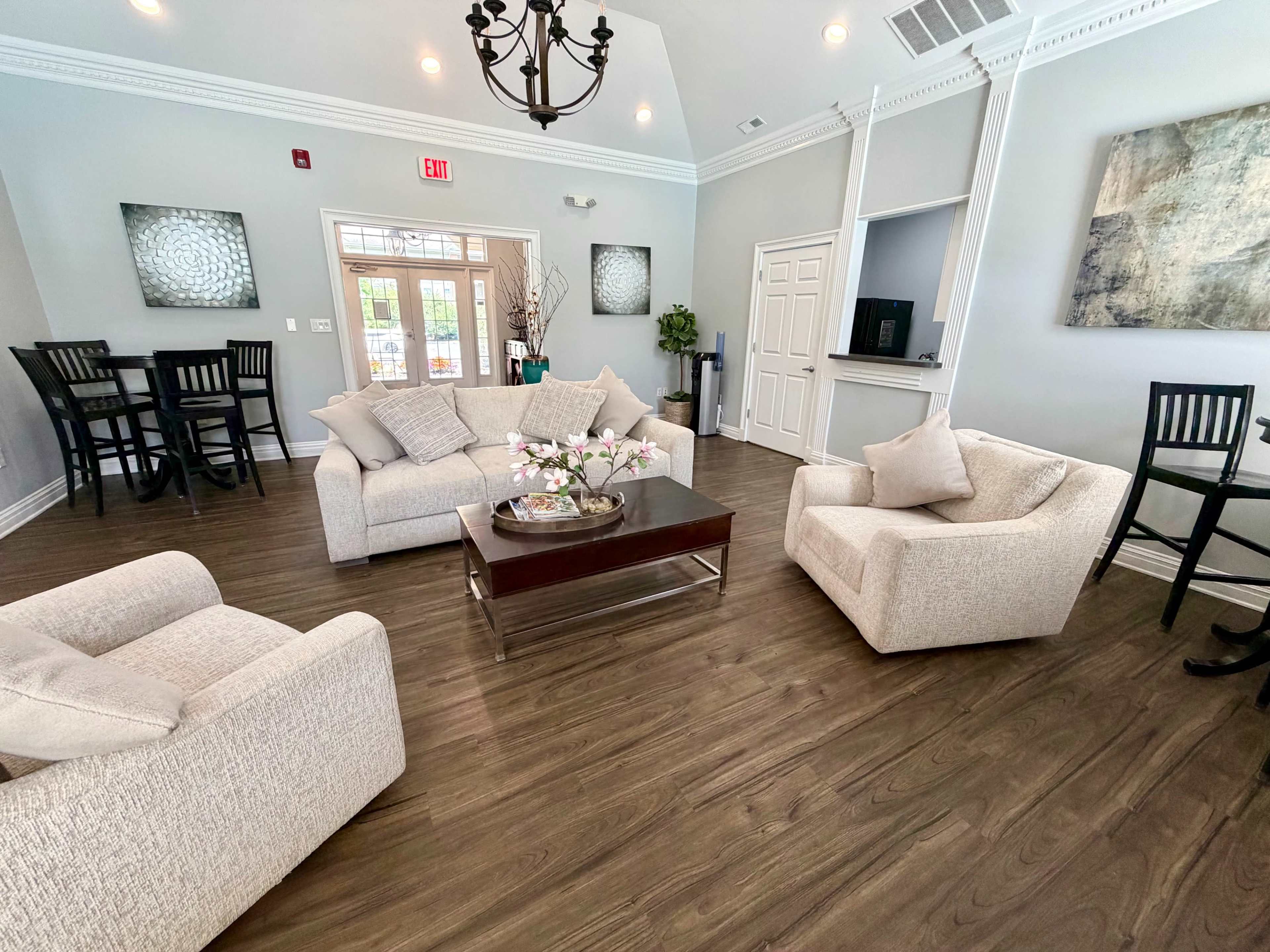 The image depicts a modern lounge area with light-colored sofas, a coffee table, and a dining table, featuring neutral decor and large windows for natural light.