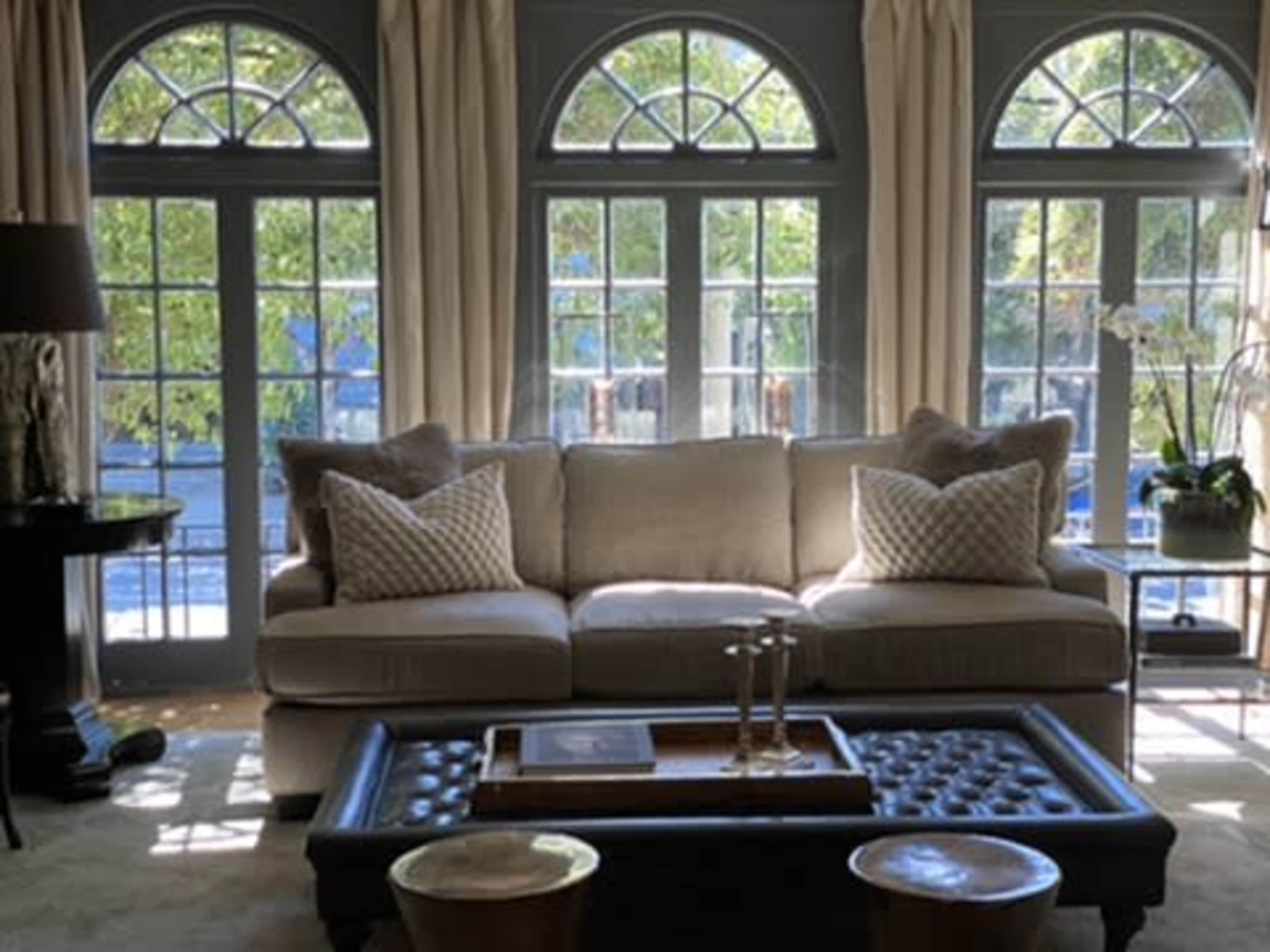A light and airy living room features a beige sofa with decorative pillows, a wooden coffee table, and large windows allowing natural light to fill the space.