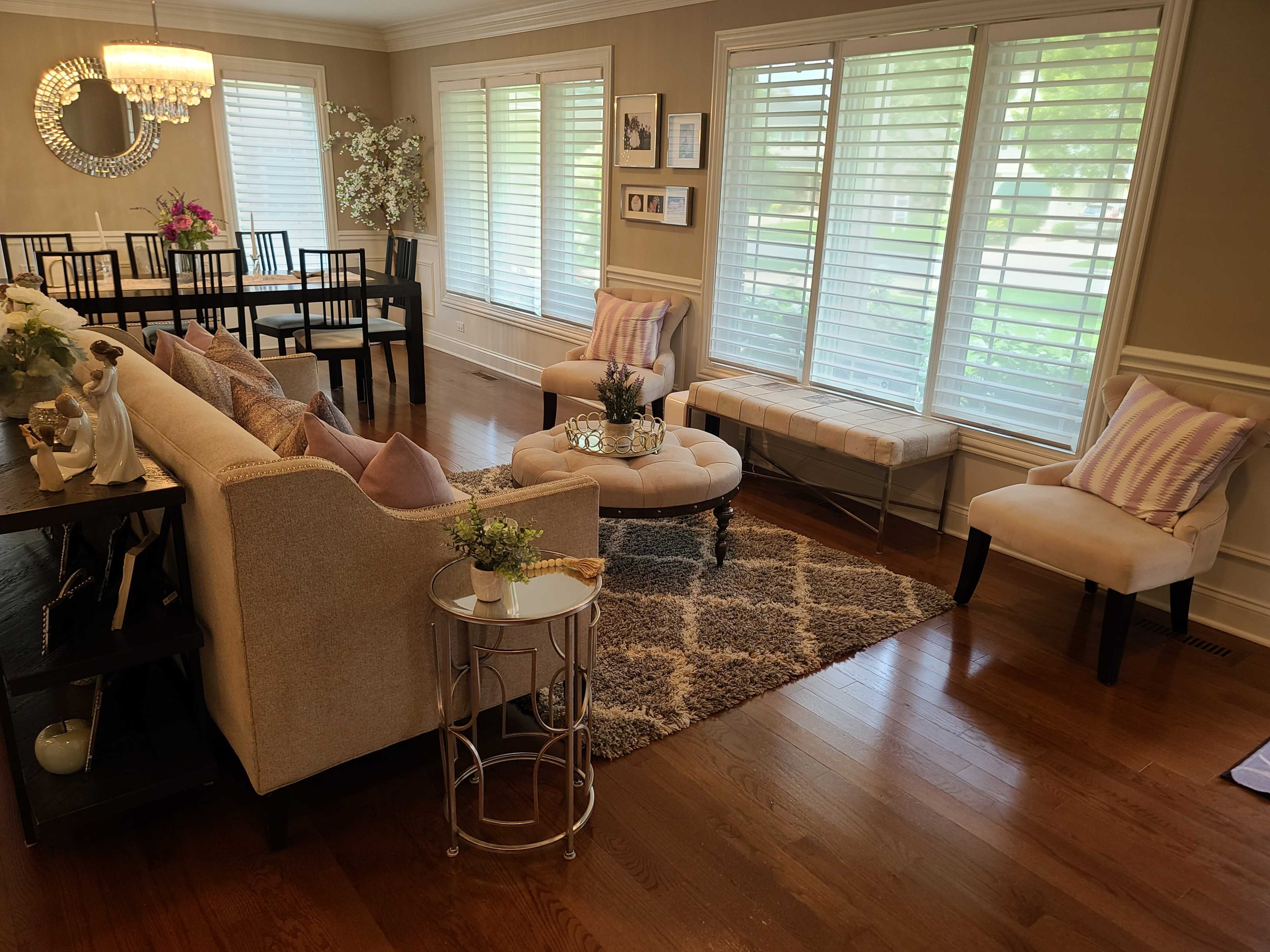 The room features a beige sofa facing a round ottoman, with a dining table set for six in the background and large windows allowing natural light to enter.