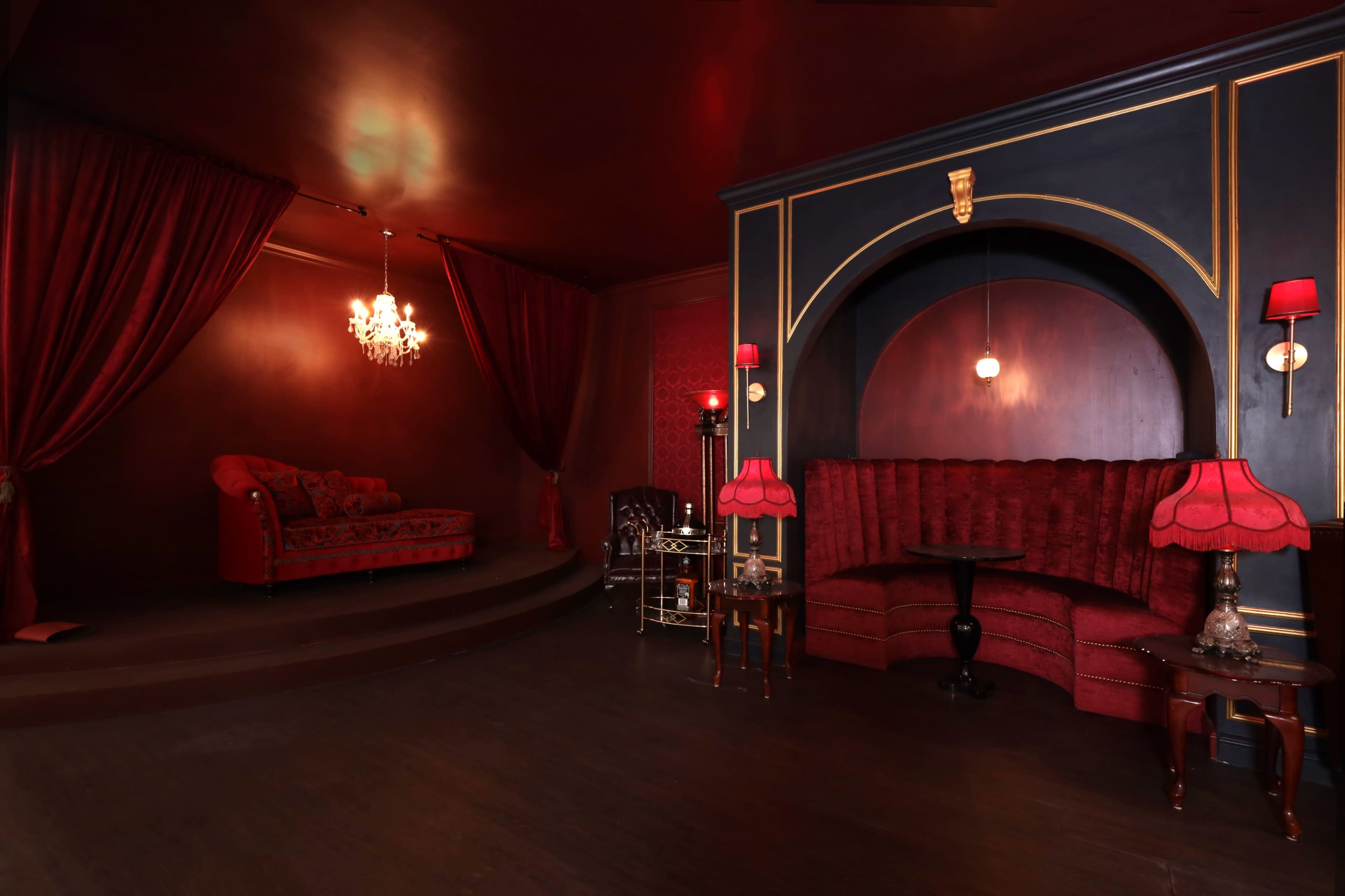 A dimly lit lounge decorated with rich red drapes, a plush red couch, and a velvet booth, all set against dark walls and accented by vintage lamps and a chandelier.