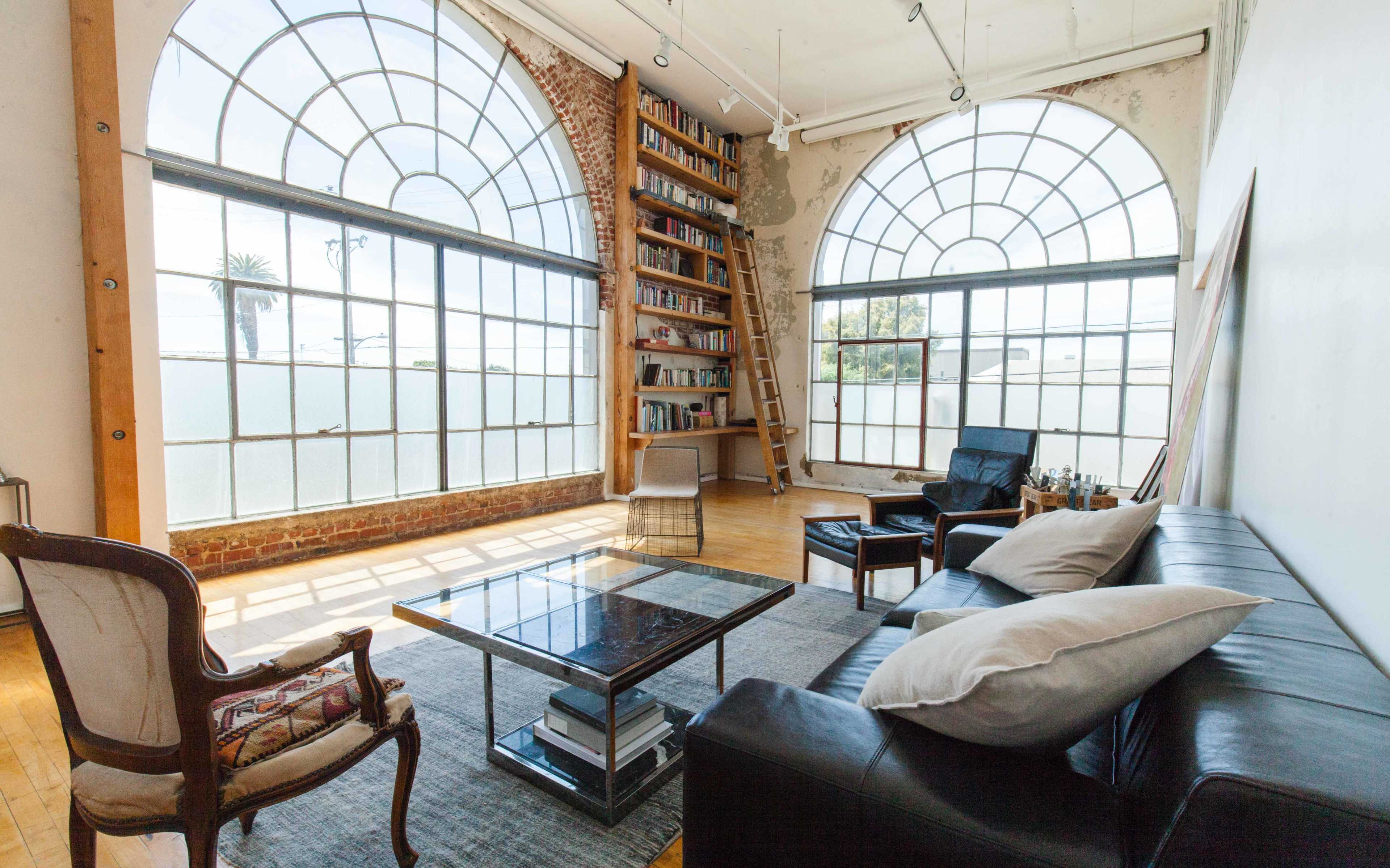 The image shows a spacious room with large arched windows, a bookshelf on a ladder, a coffee table, and a seating area with black leather chairs and a patterned armchair.