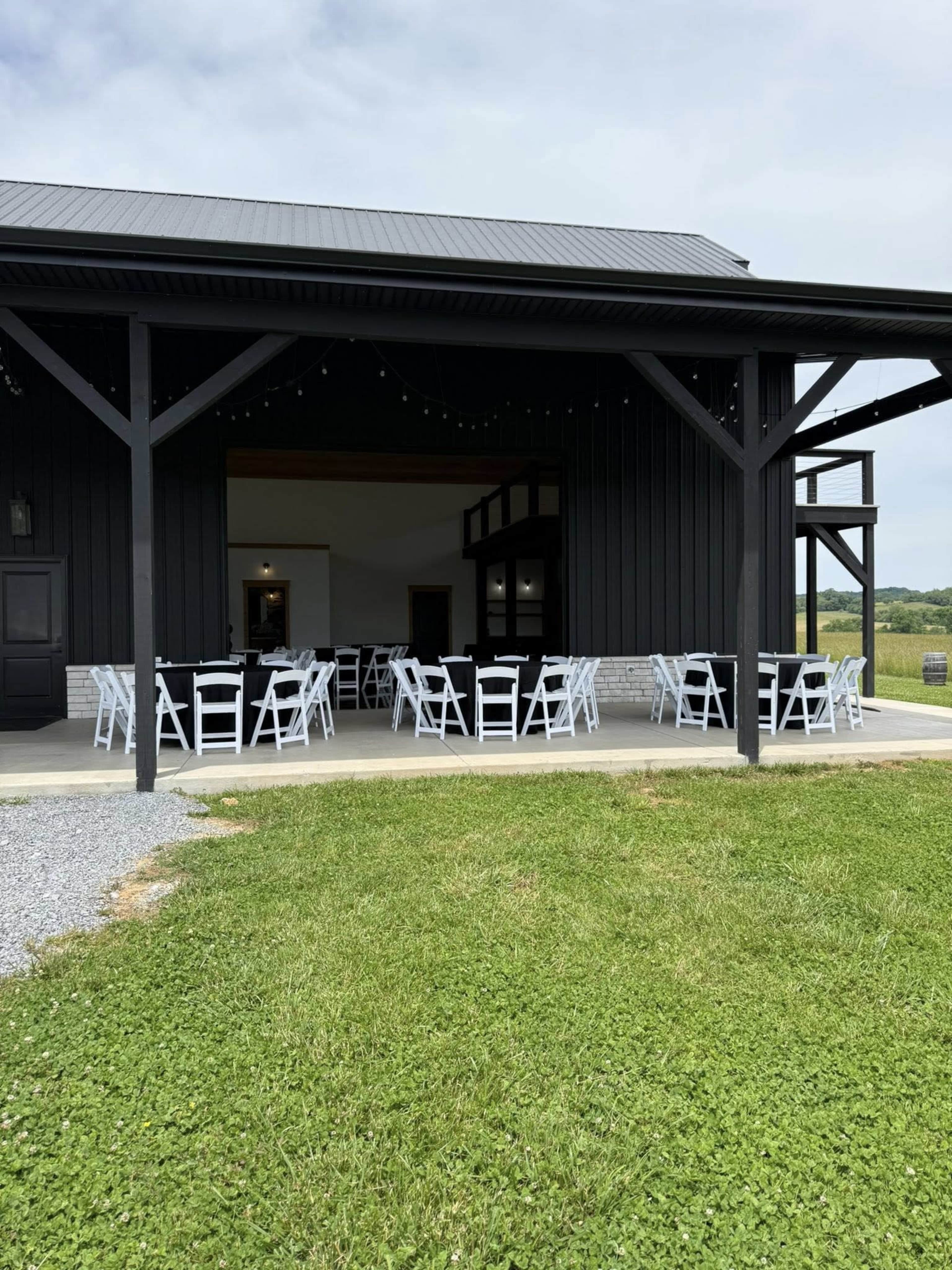 The image shows a venue with a covered outdoor seating area featuring several white tables and chairs on a grassy lawn.