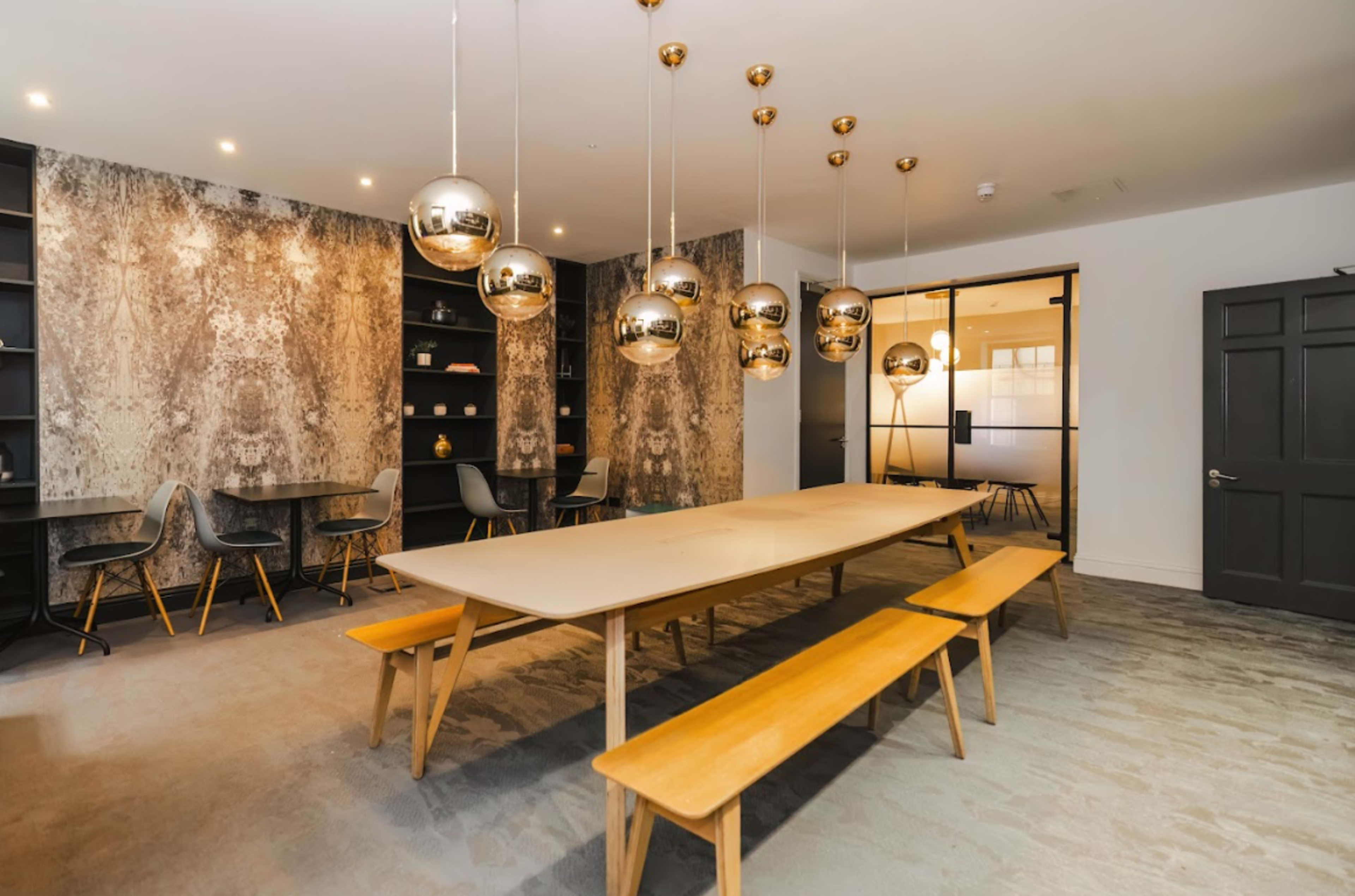 A light-filled dining area features a long wooden table with benches and modern pendant lights hanging above, set against a textured wall.