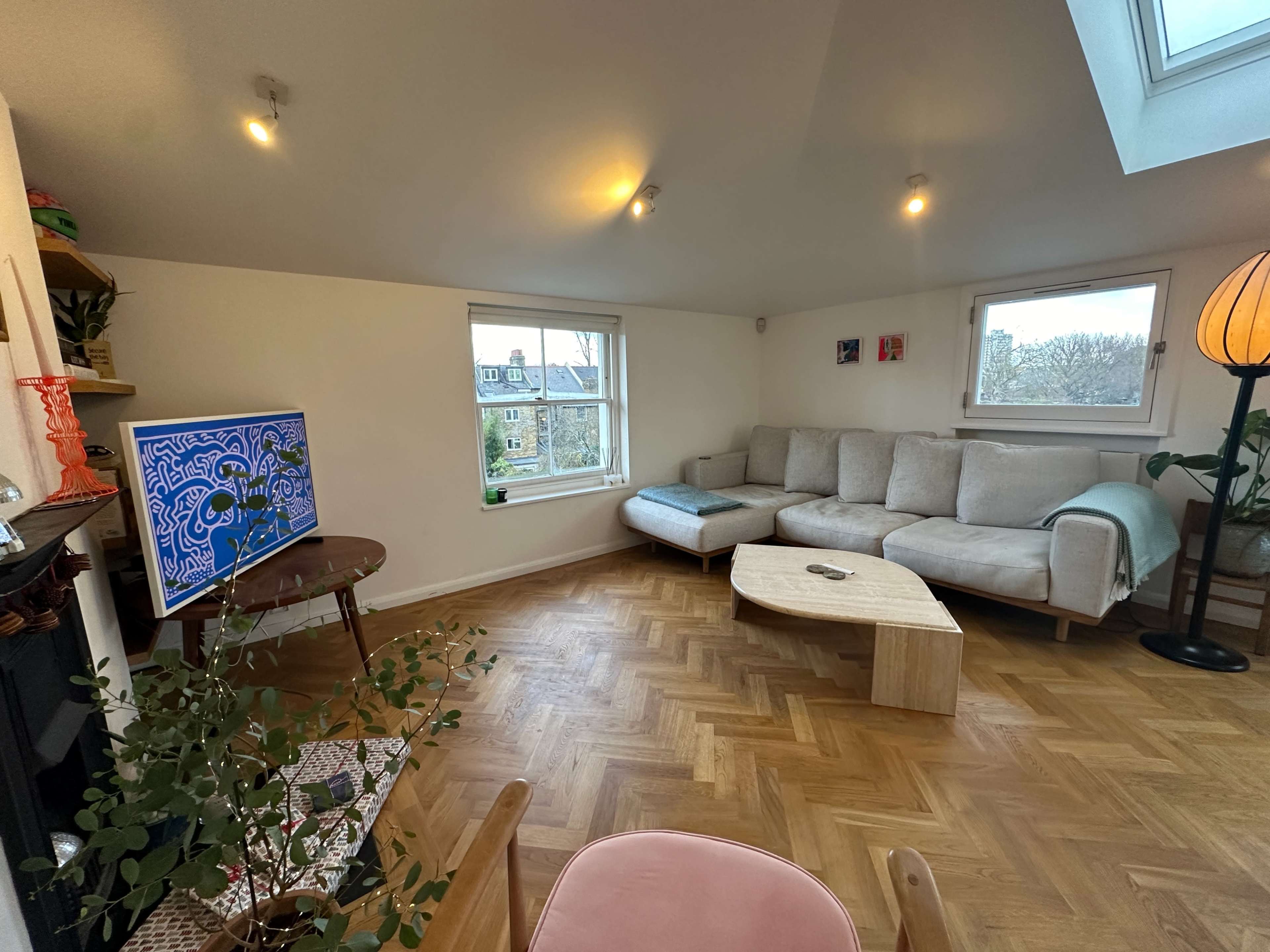 The image shows a cozy living room with a sectional sofa, a small coffee table, a television on a stand, and a large window letting in natural light.