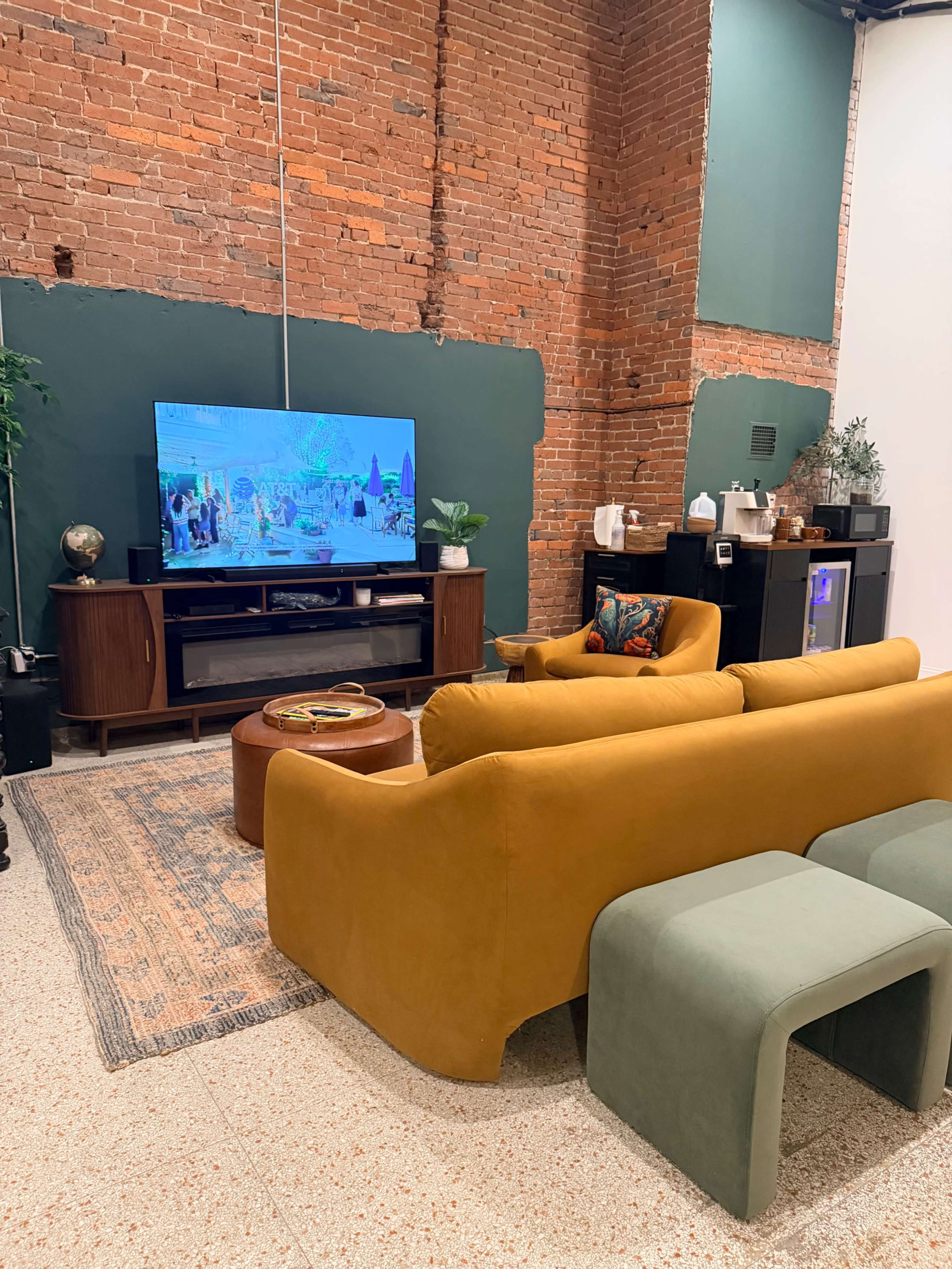 The image shows a modern living room featuring a yellow sofa, a brown coffee table, a large TV mounted on a brick wall, and green accent walls.
