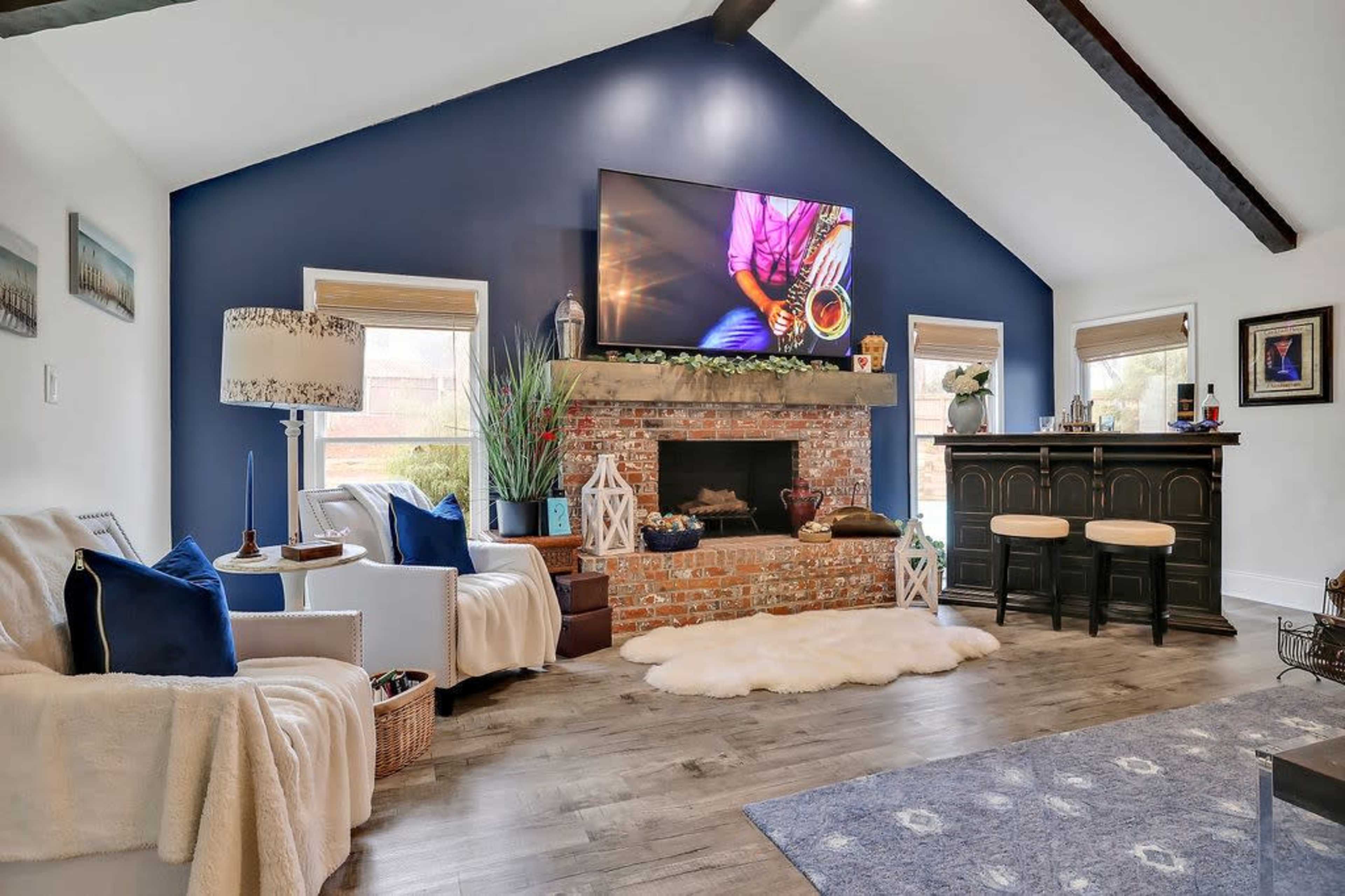 The living room features a brick fireplace with a mounted TV above it, flanked by two windows and furnished with a sofa, a lamp, and a bar area.