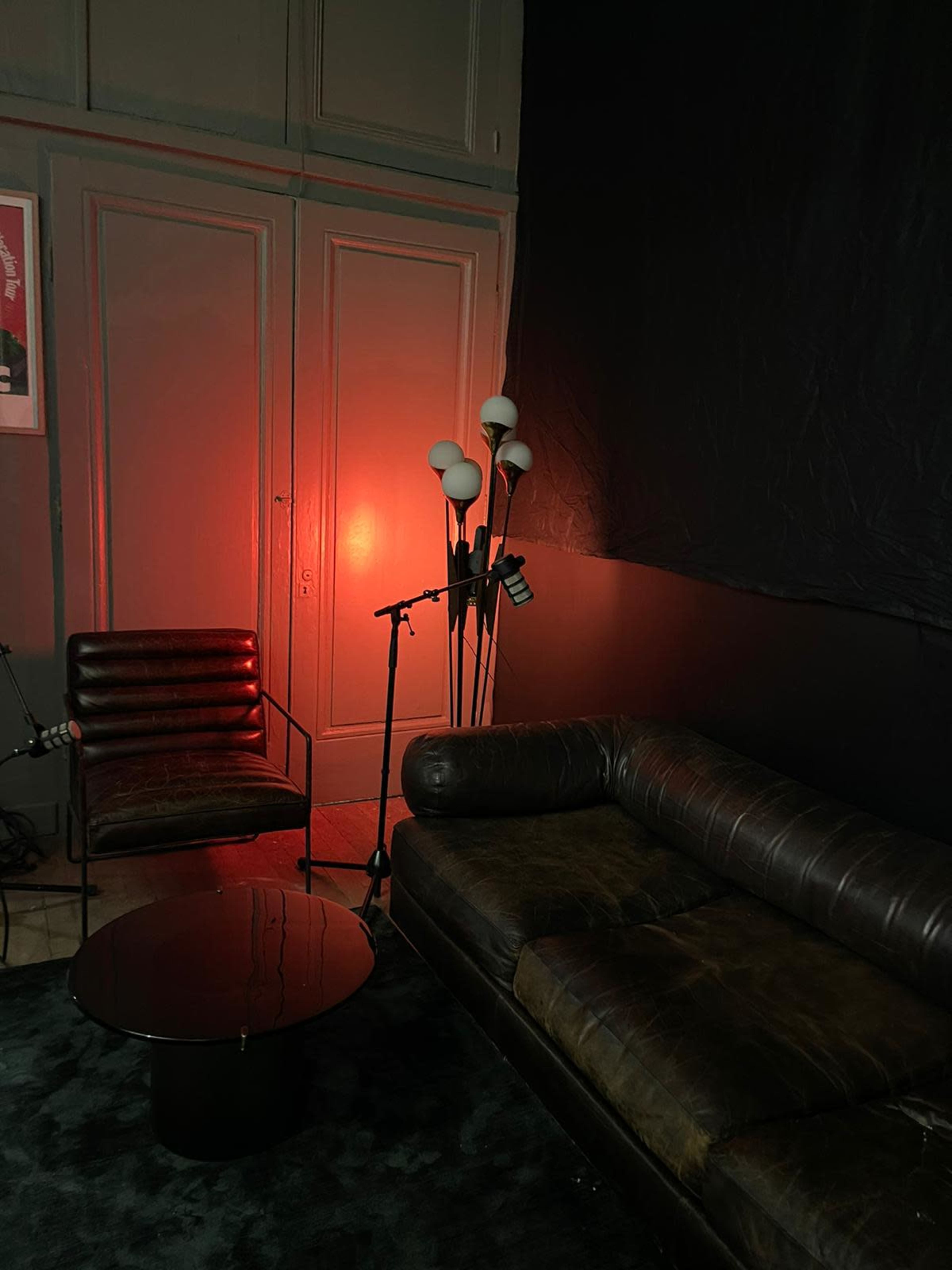 A dimly lit room features a leather sofa, a metal side table, an armchair, and a floor lamp with multiple bulbs.