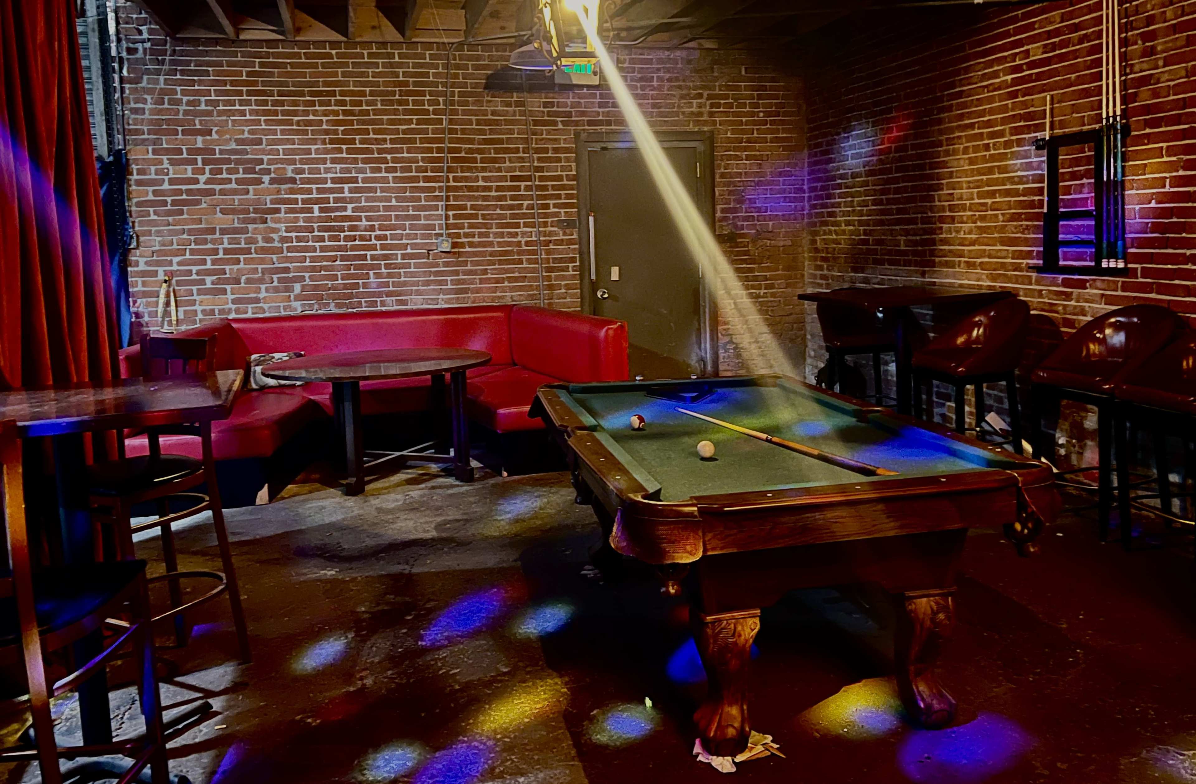 A dimly lit room features a pool table with three balls and a red sofa against a brick wall.